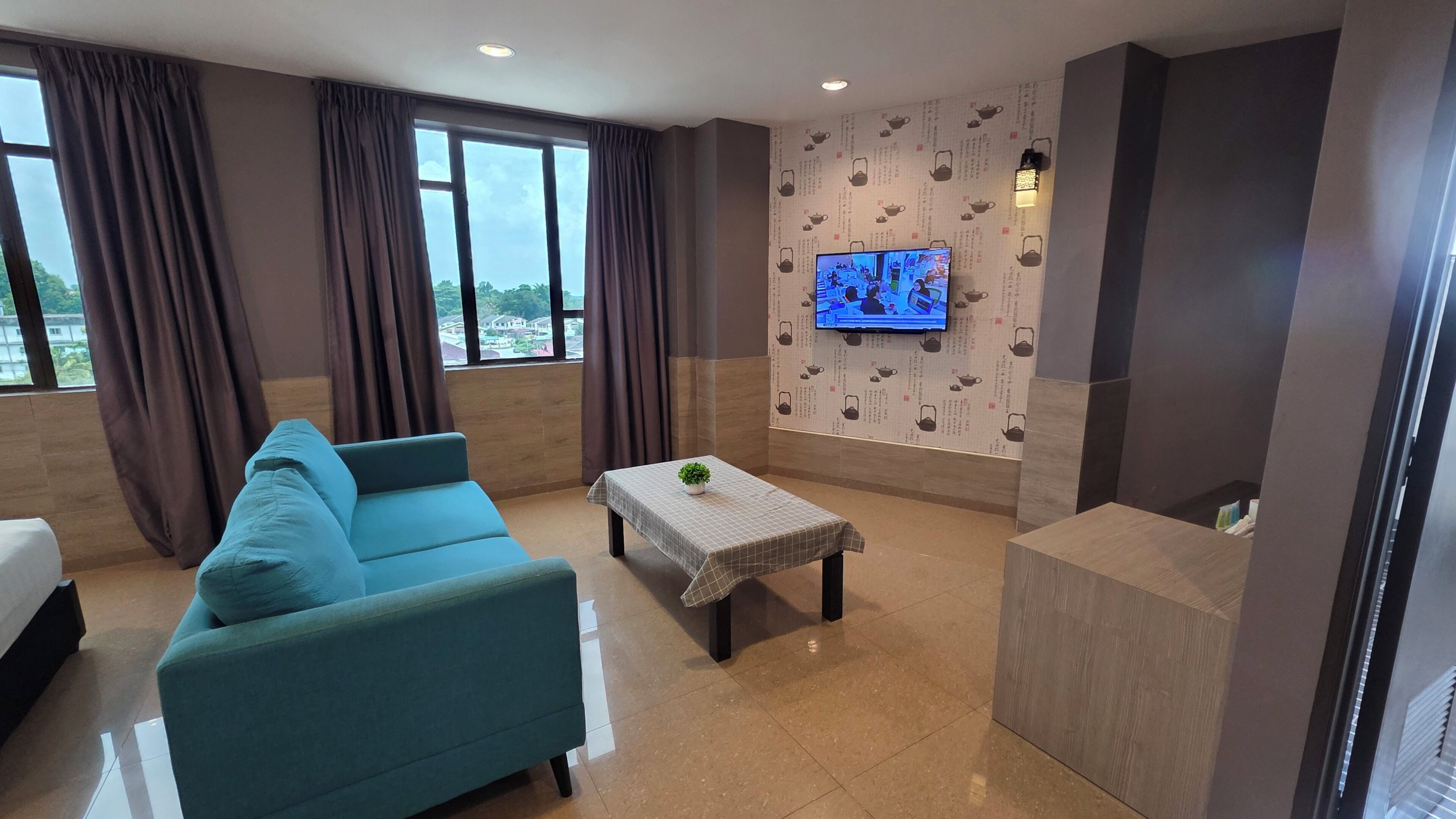 A modern living room with a teal sofa, a small table with a plant, a flat-screen TV on the wall, and large windows with curtains.
