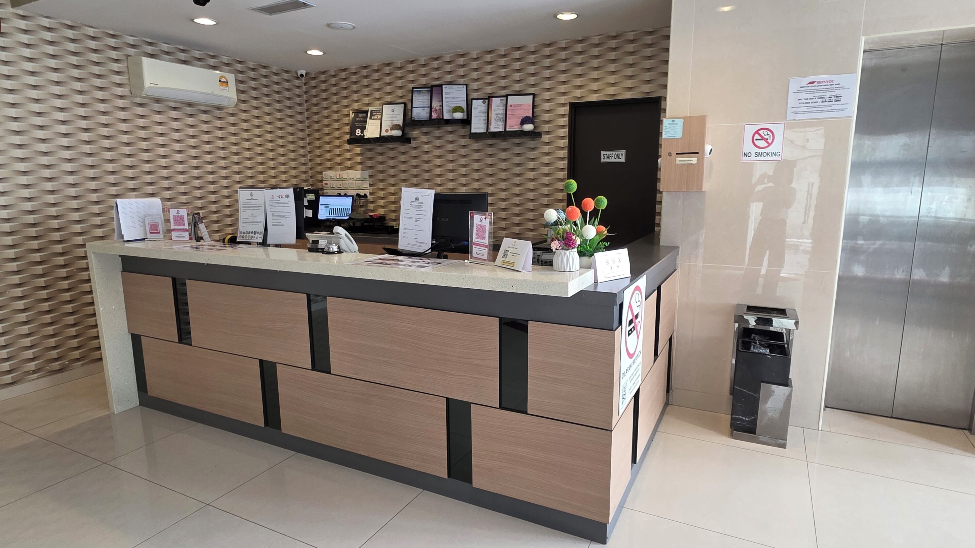 Bancada de recepção com computador, telefone e folhetos informativos. Placas indicam 'Apenas para funcionários' e 'Proibido fumar'.