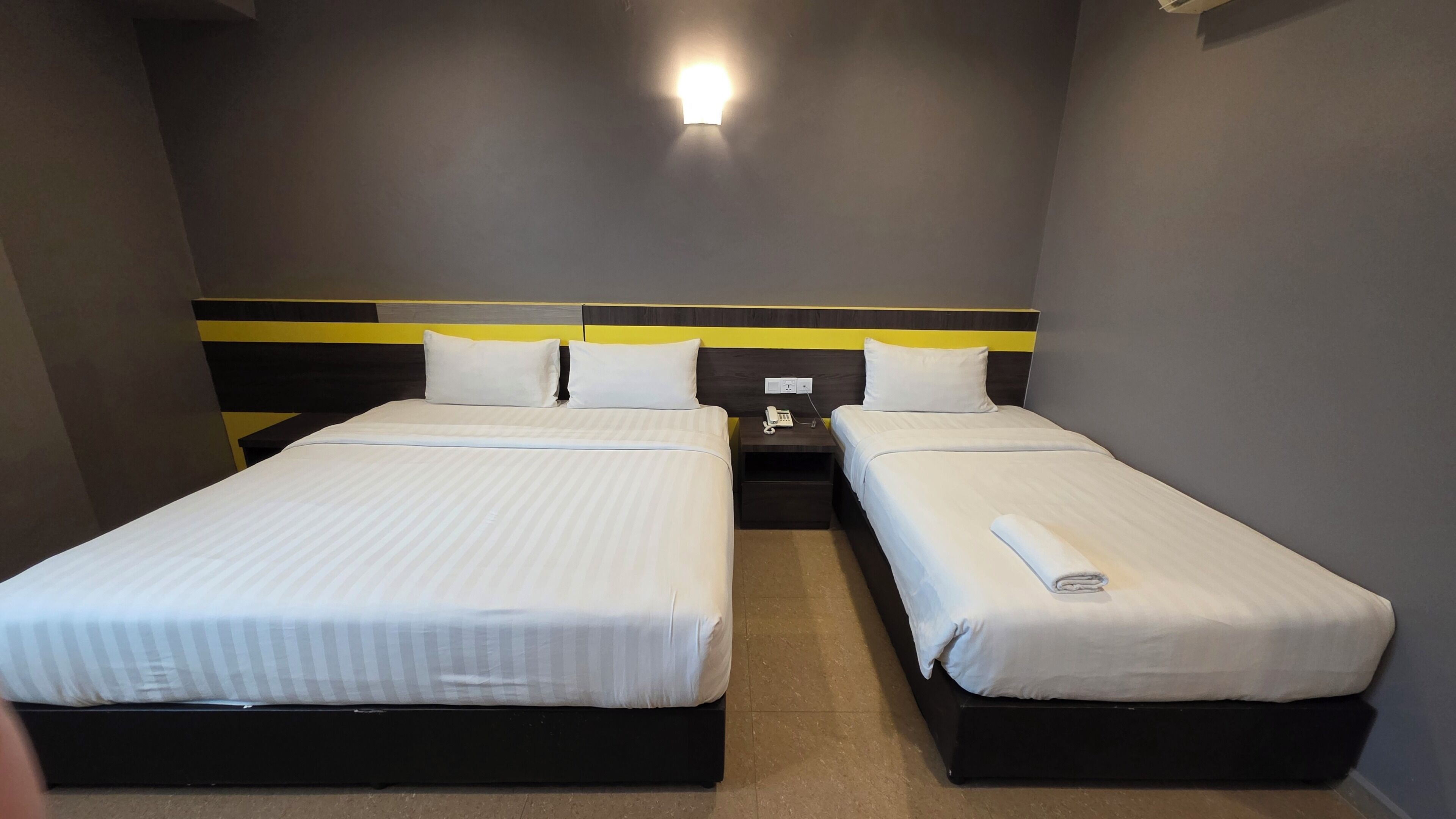 A hotel room with two single beds, a nightstand with a phone, and a wall-mounted light.