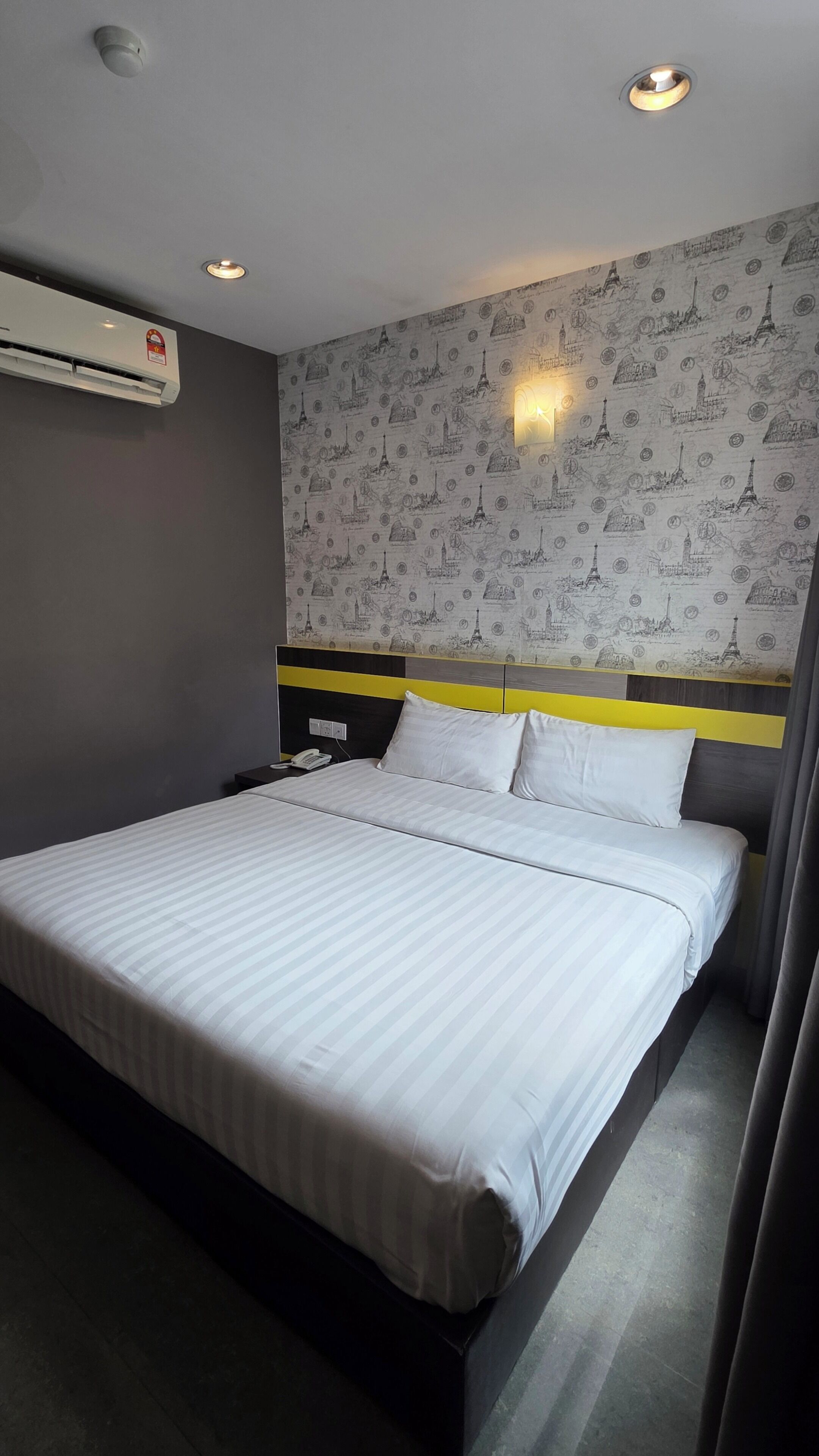 A hotel room with a bed, a wall-mounted air conditioner, a nightstand with a phone, and a wall with a pattern of famous landmarks.