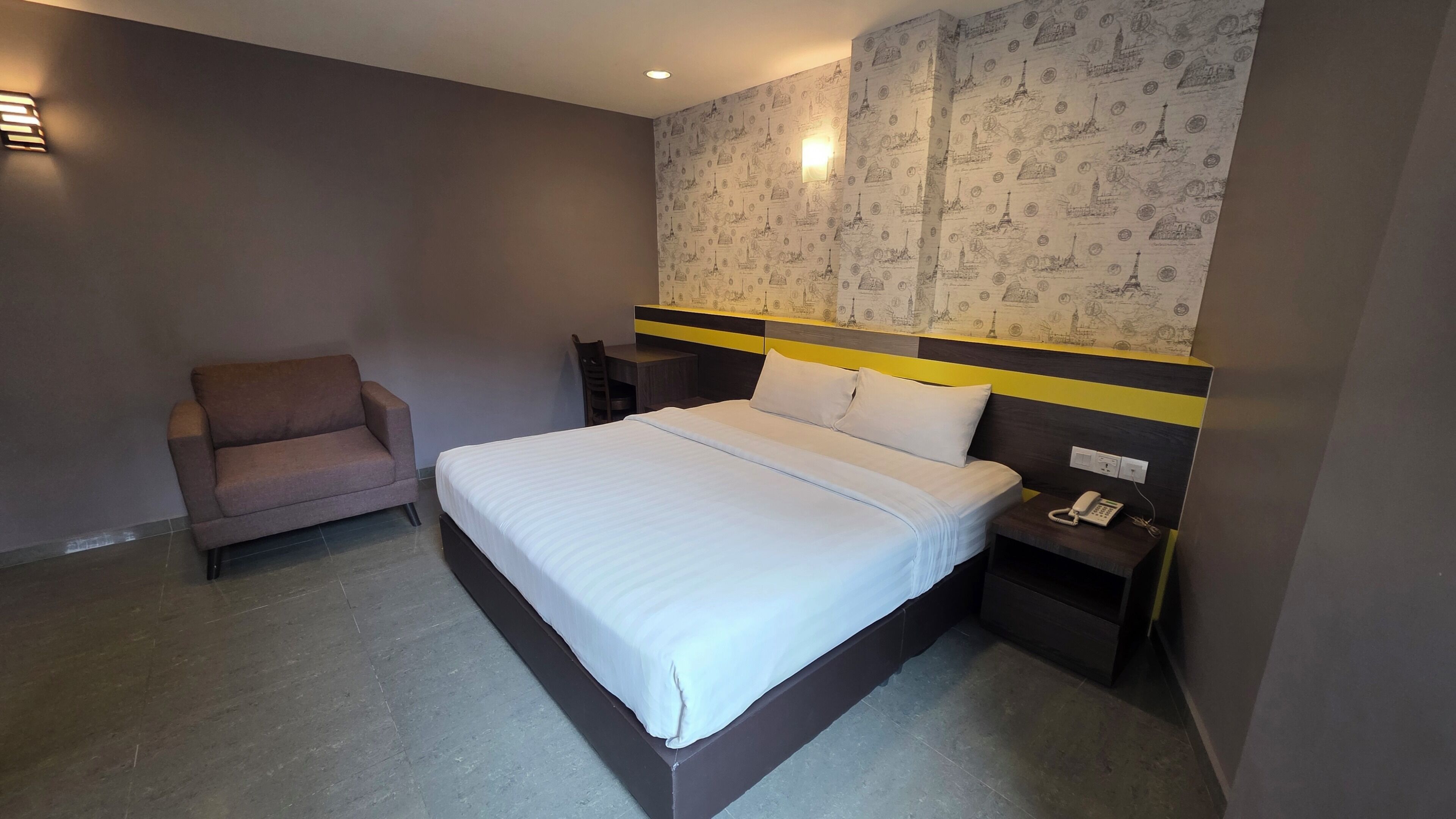 Une chambre d’hôtel comprenant un lit, une chaise, une table de chevet avec un téléphone et un mur décoré d’un motif représentant un paysage urbain.