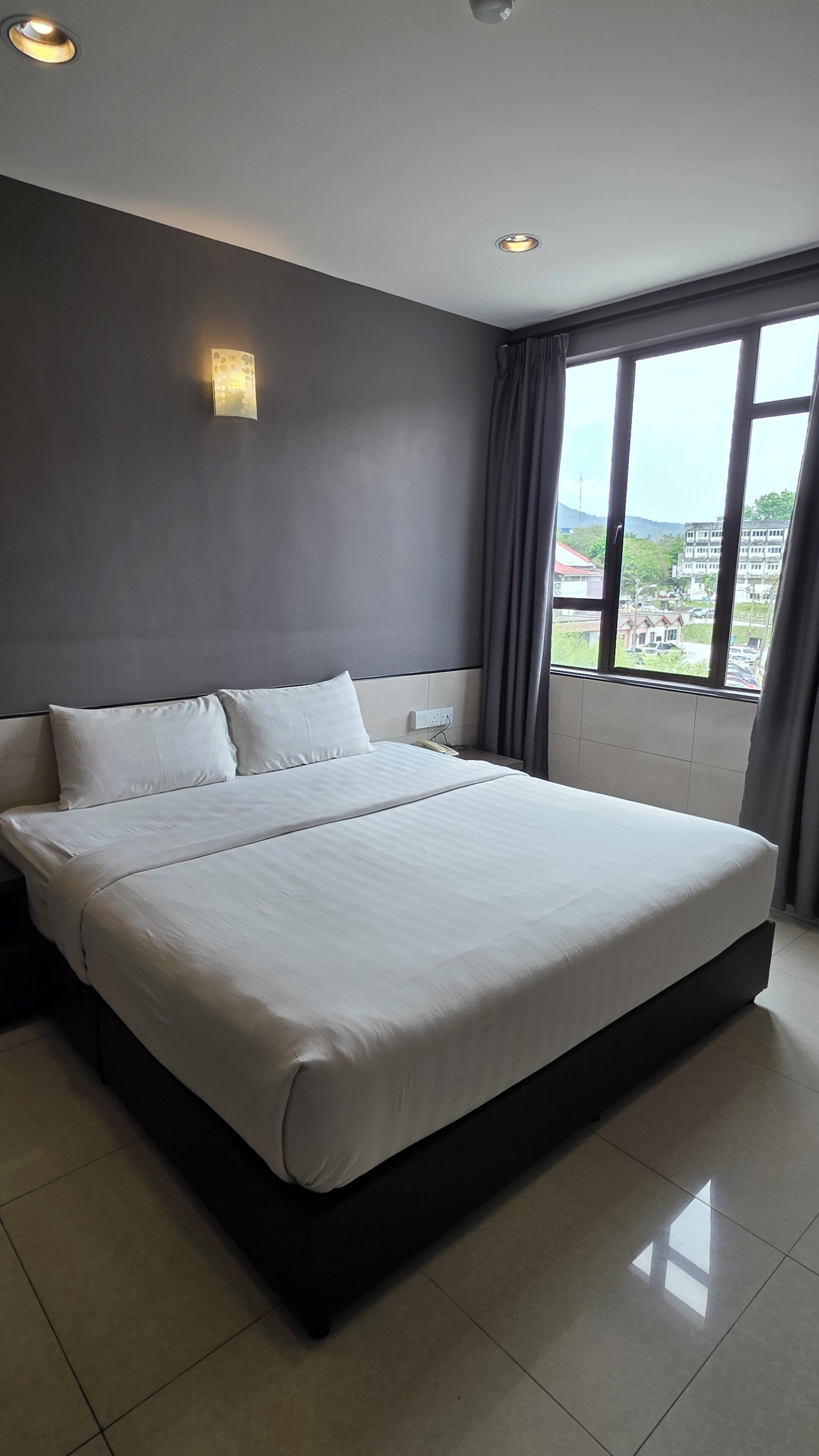 A modern hotel room with a large bed, a bedside table, a wall-mounted light, and a window with a view of the city.