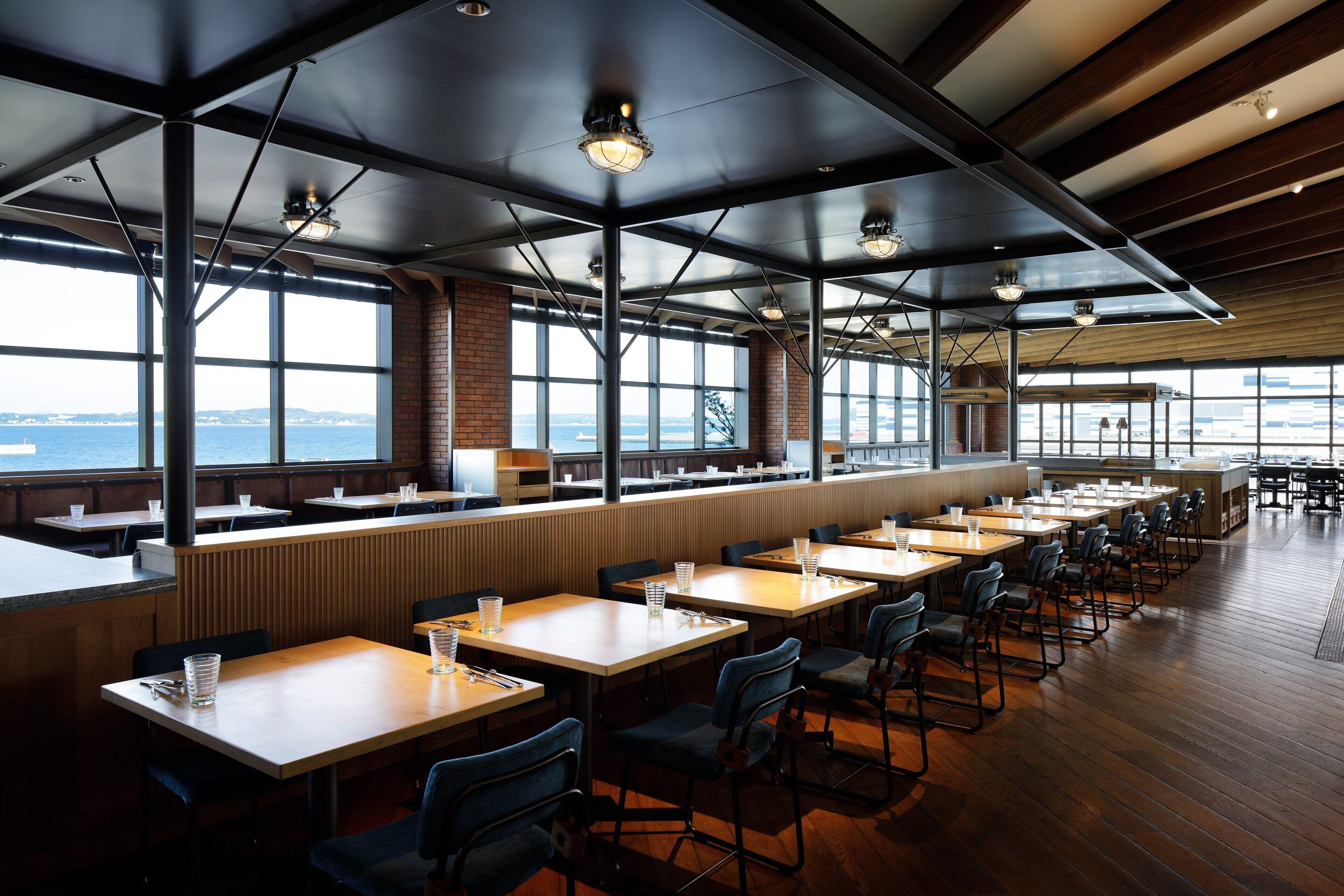 En rymlig restaurang med högt i tak, stora fönster och utsikt över havet. Borden är snyggt uppställda med stolar.