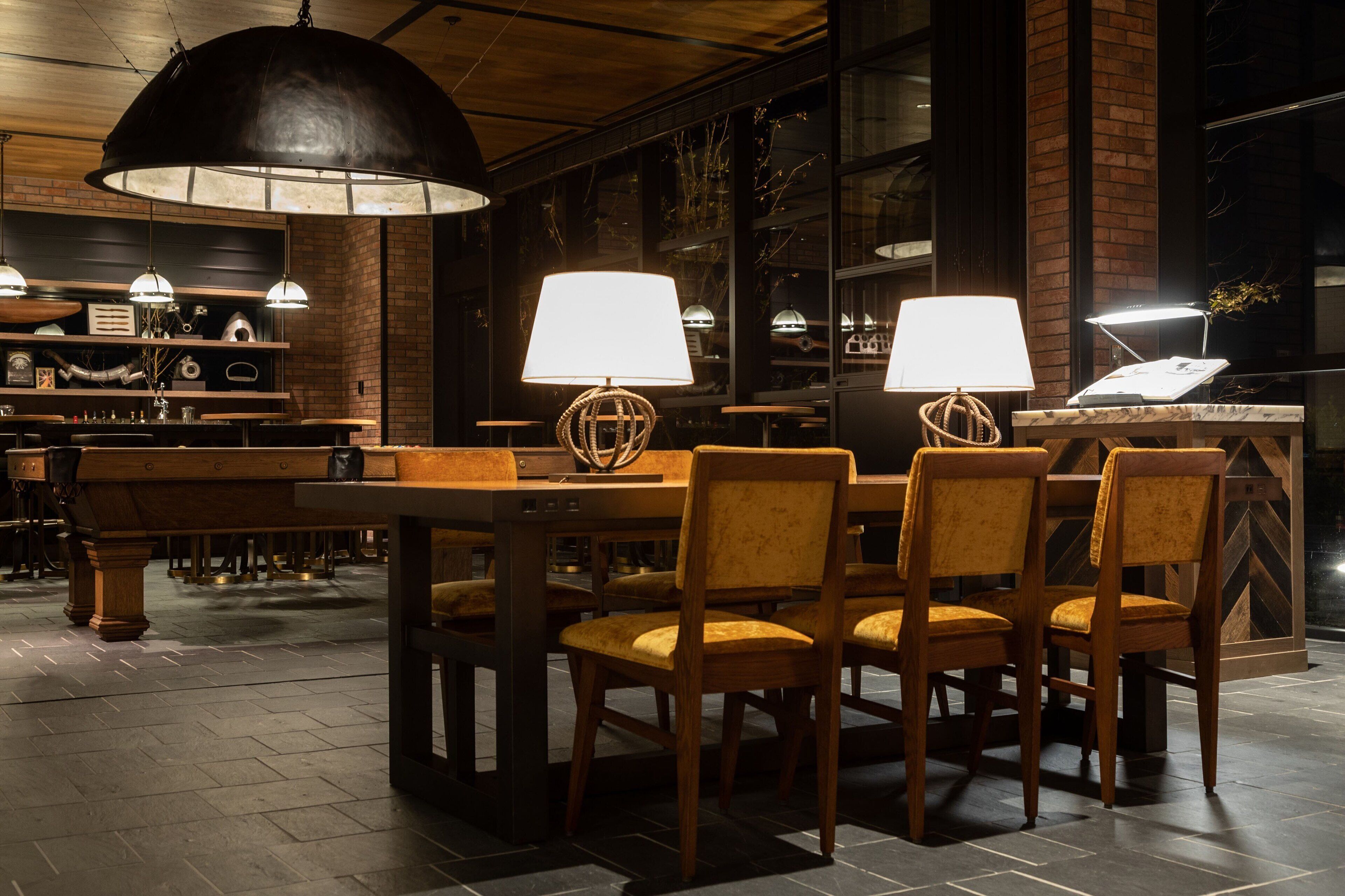 A restaurant with a brick wall, wooden tables, and chairs. There are pendant lights and a large black lamp over the tables.