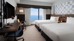 Een hotelkamer met twee bedden, een bureau met stoel, een televisie en een groot raam met uitzicht op de zee.