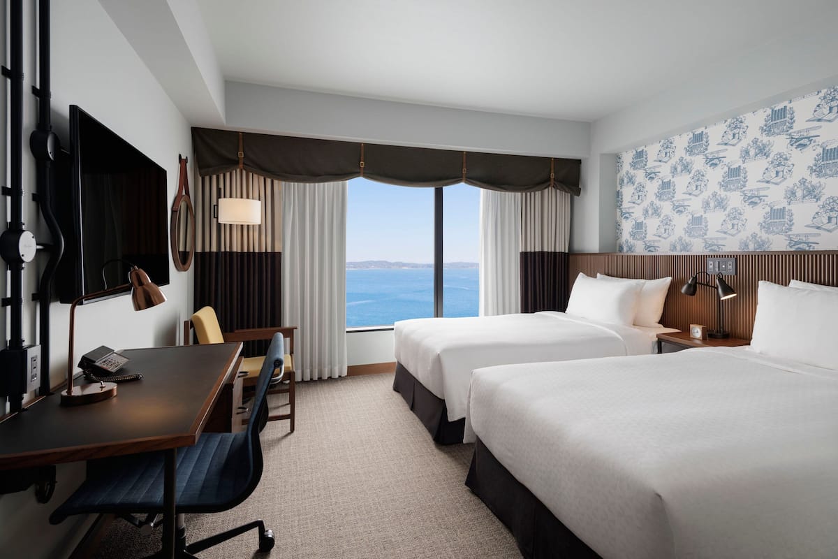 Une chambre d’hôtel avec deux lits, un bureau avec une chaise, une télévision et une grande fenêtre donnant sur la mer.