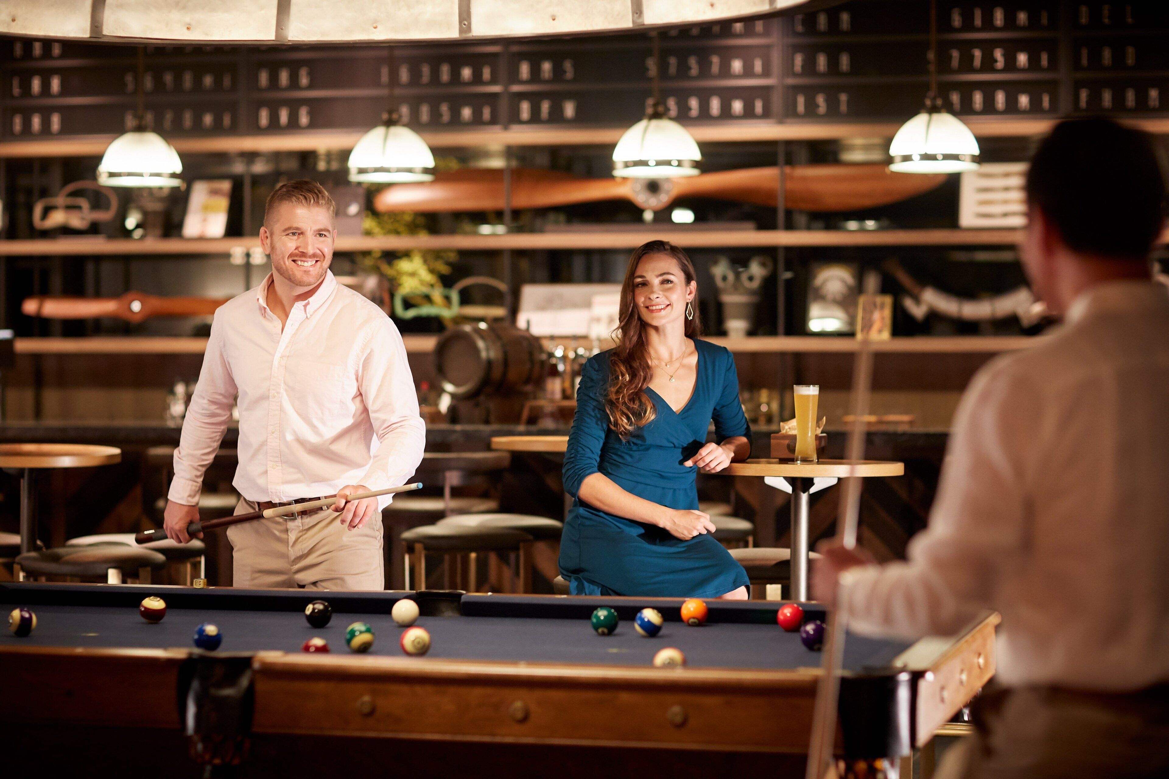 A billiards table with balls and cues, a woman in a blue dress, and a man in a light pink shirt in a room with wooden shelves and hanging lights.