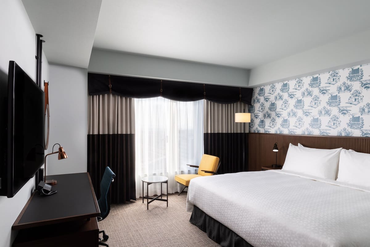 Une chambre d’hôtel avec un lit, un bureau avec une lampe, une chaise, une télévision et une fenêtre avec des rideaux.