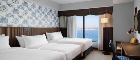 A hotel room with two beds, a desk, and a view of the sea.