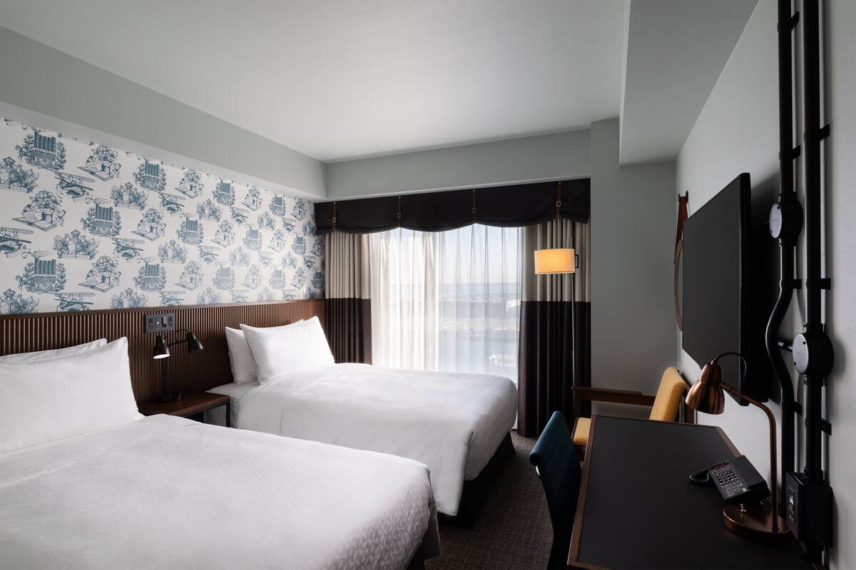 Une chambre d’hôtel avec deux lits, un bureau, une chaise, une télévision et une fenêtre avec des rideaux.