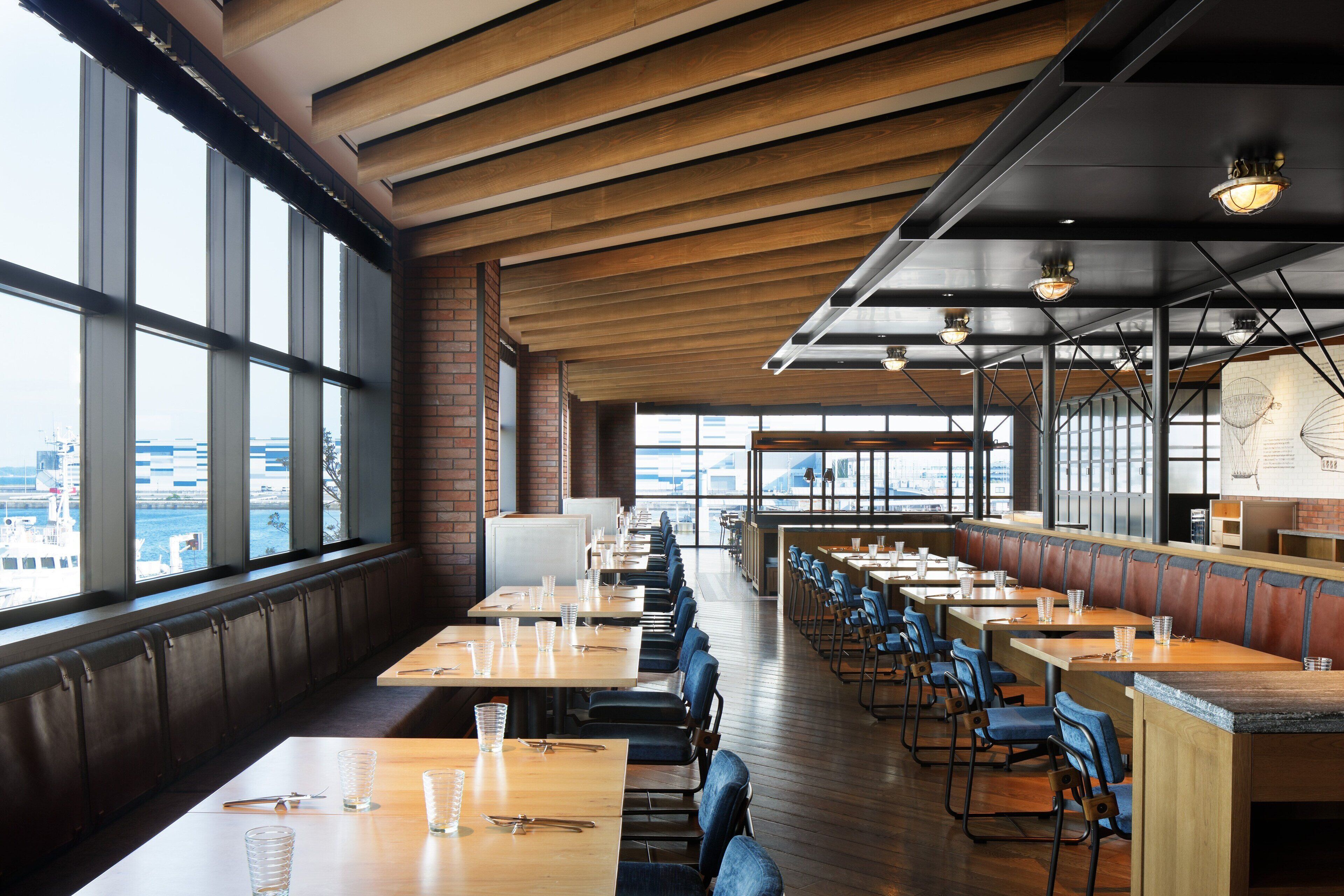 A restaurant with long wooden tables and blue chairs, large windows, and a view of the city.