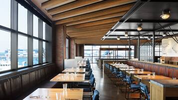A restaurant with long wooden tables and blue chairs, large windows, and a view of the city.