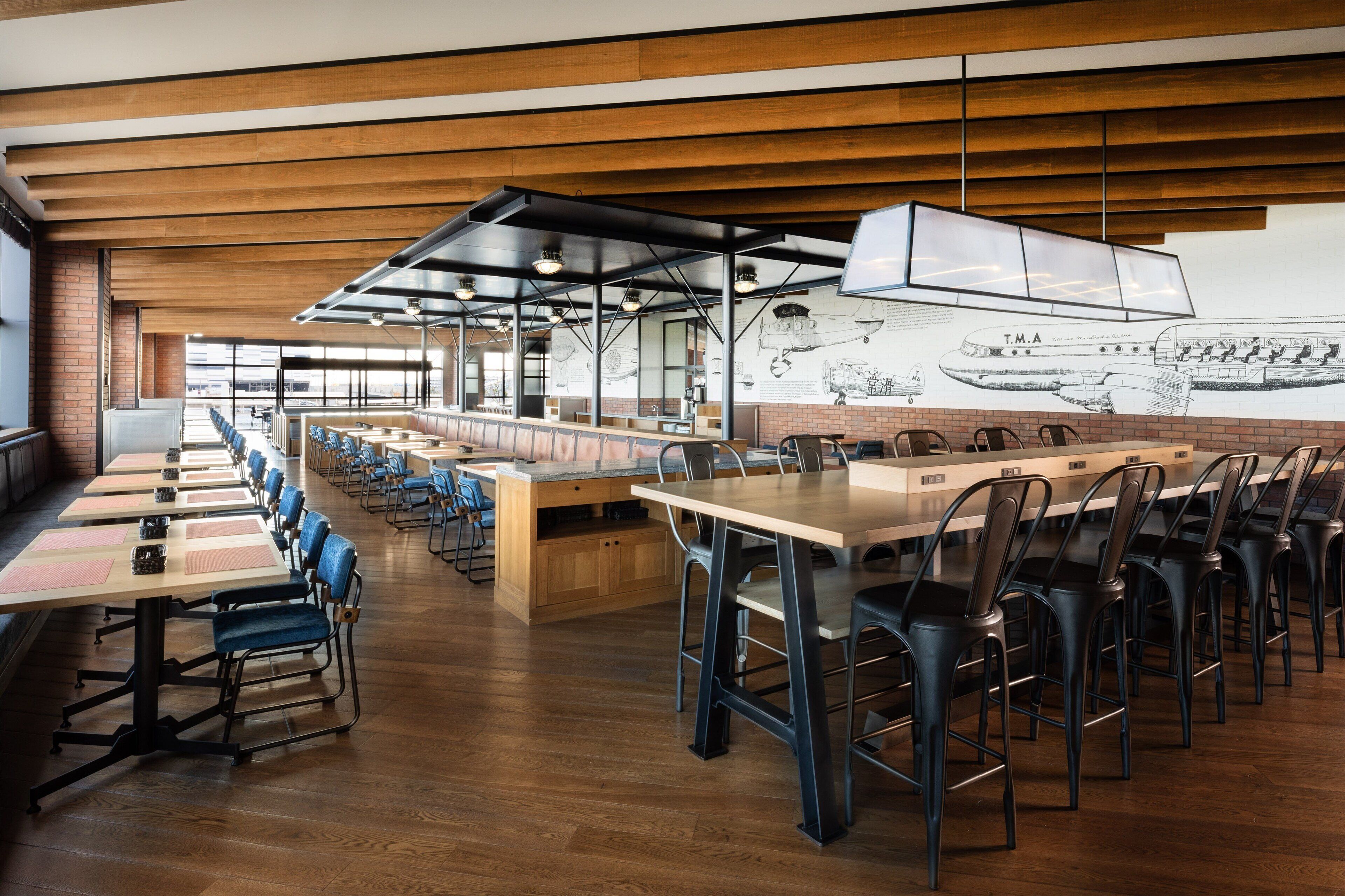 A spacious restaurant with long communal tables and blue chairs, wooden floors, and a large mural of an airplane on the wall.