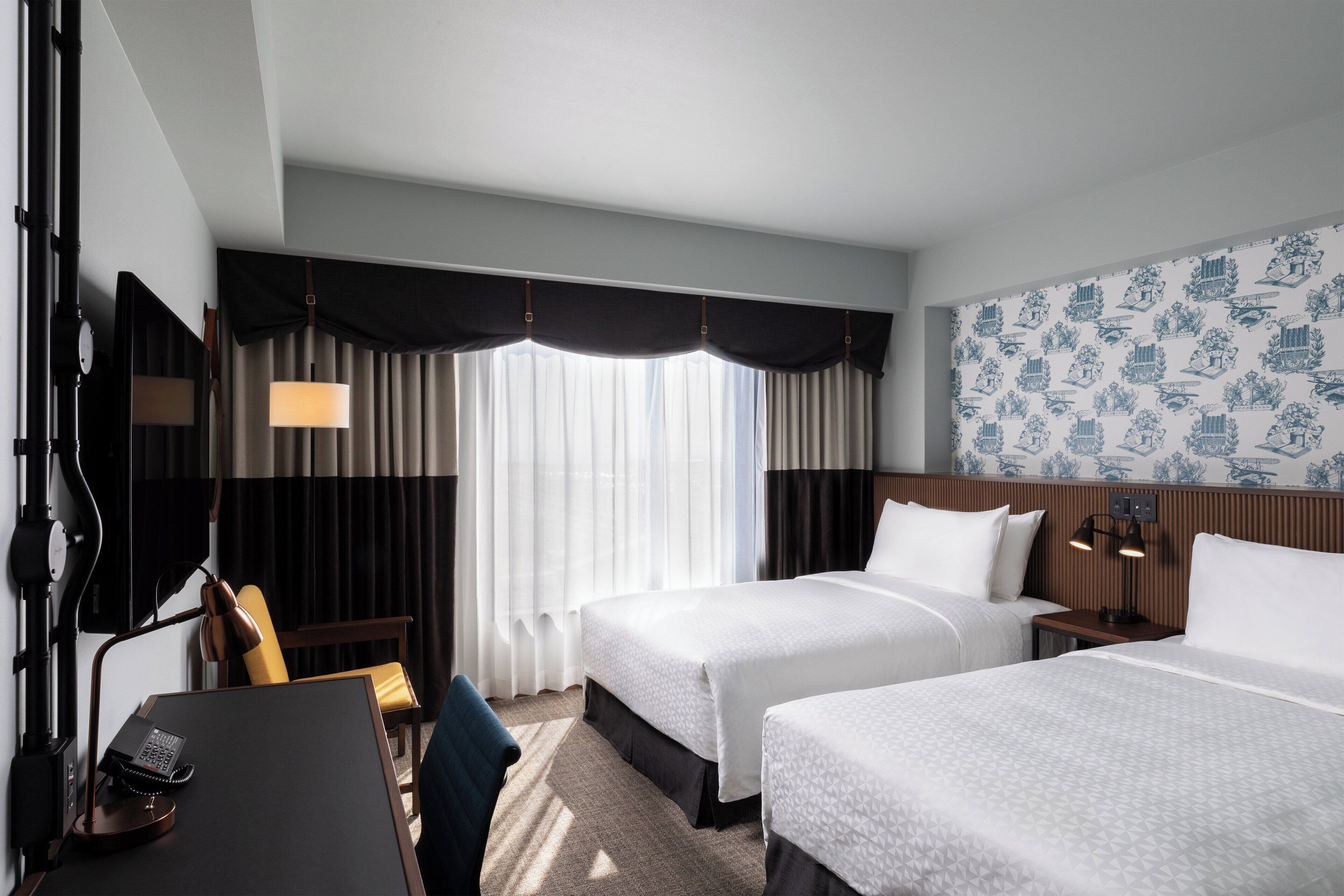 A hotel room with two beds, a desk with a telephone, a chair, a lamp, and a large window with curtains.