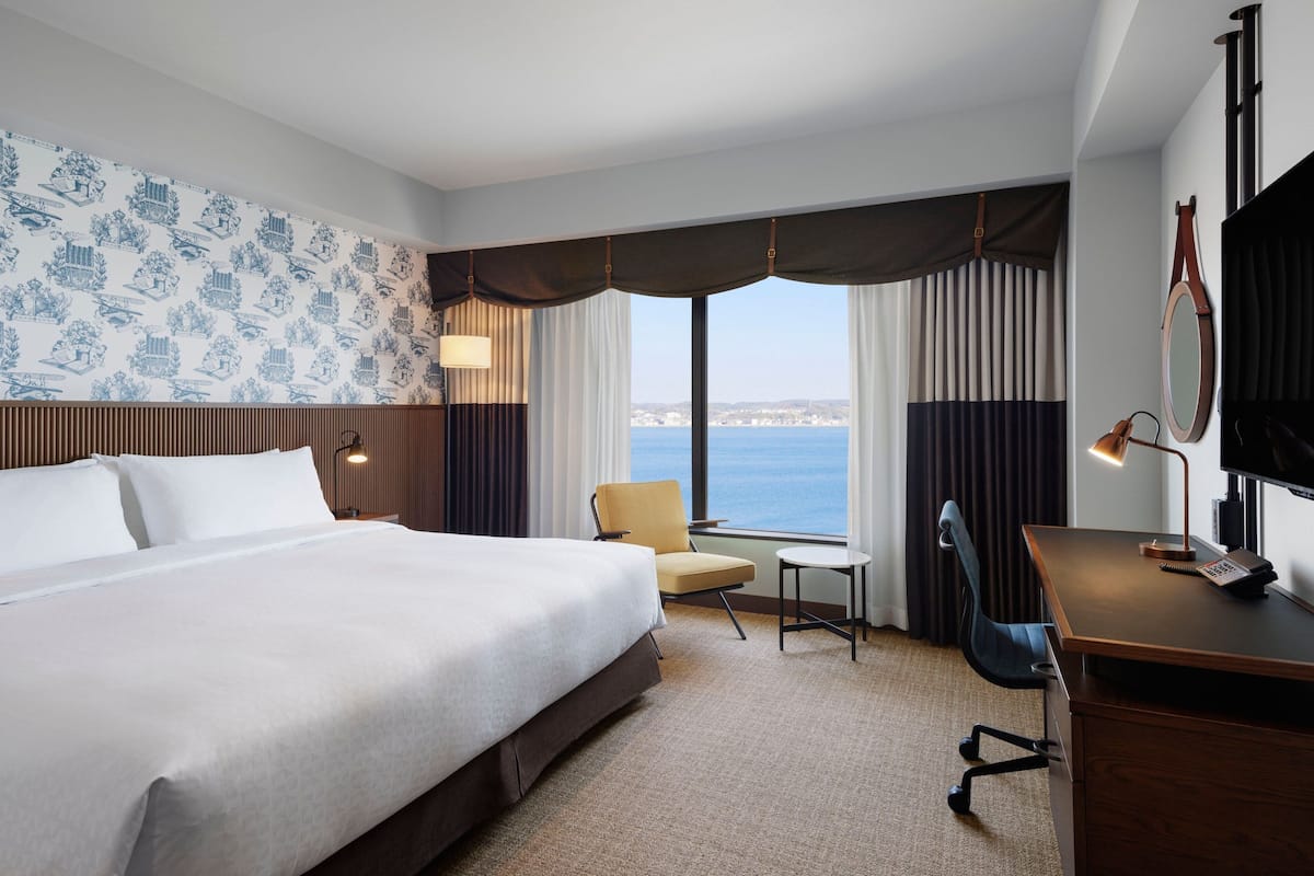 Une chambre d’hôtel avec un grand lit, un bureau, une chaise, une lampe et une vue sur la mer.