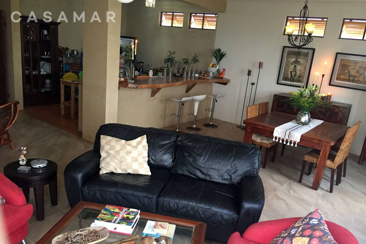 A living room with a black leather sofa, a coffee table with books and a decorative bowl, a dining area with a wooden table and chairs, and a kitchen area with a counter and plants.