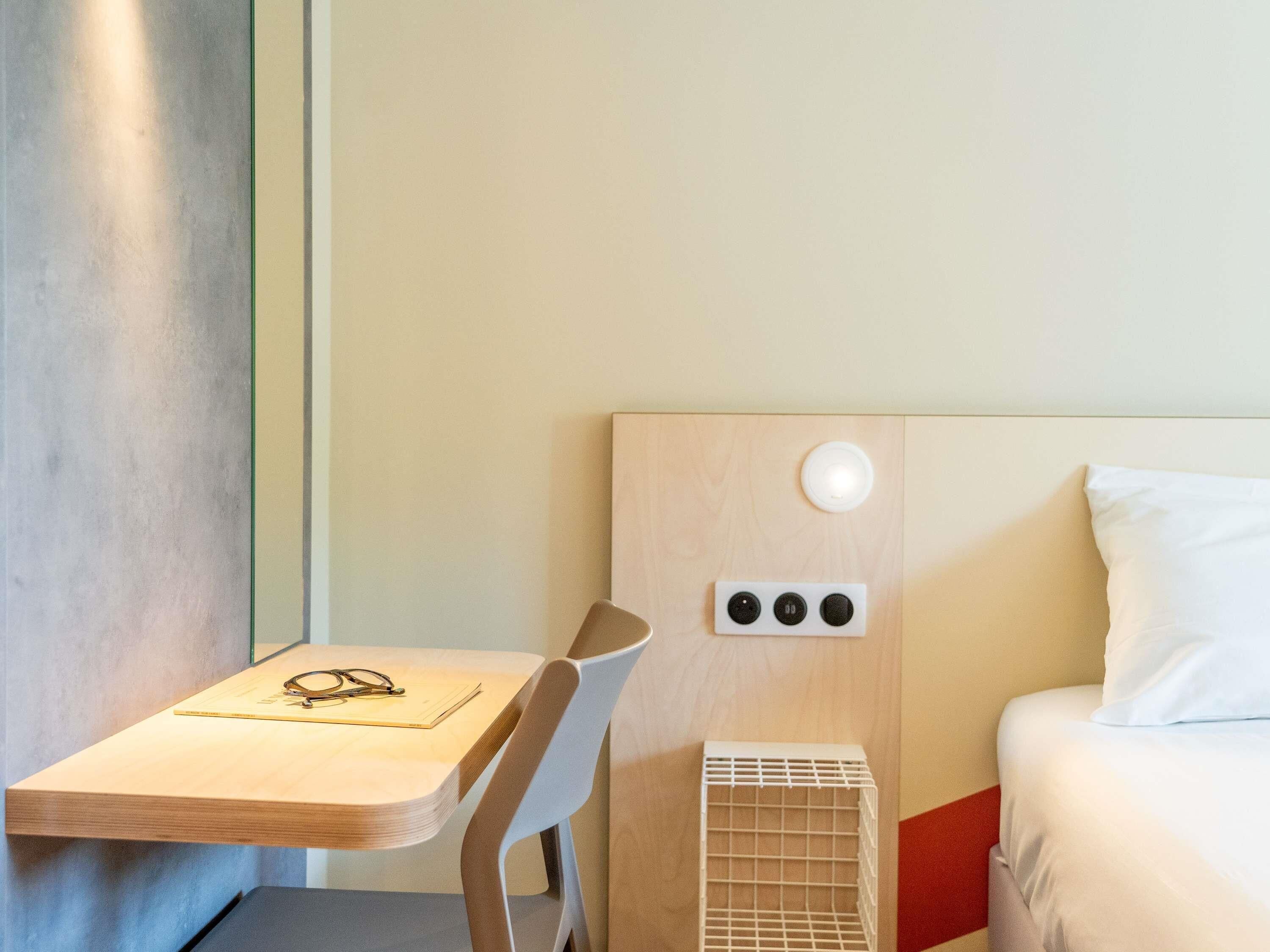 Une chambre d’hôtel avec un lit, une table de chevet avec des verres et un classeur, une chaise, une prise électrique fixée au mur et une petite grille d’aération blanche.