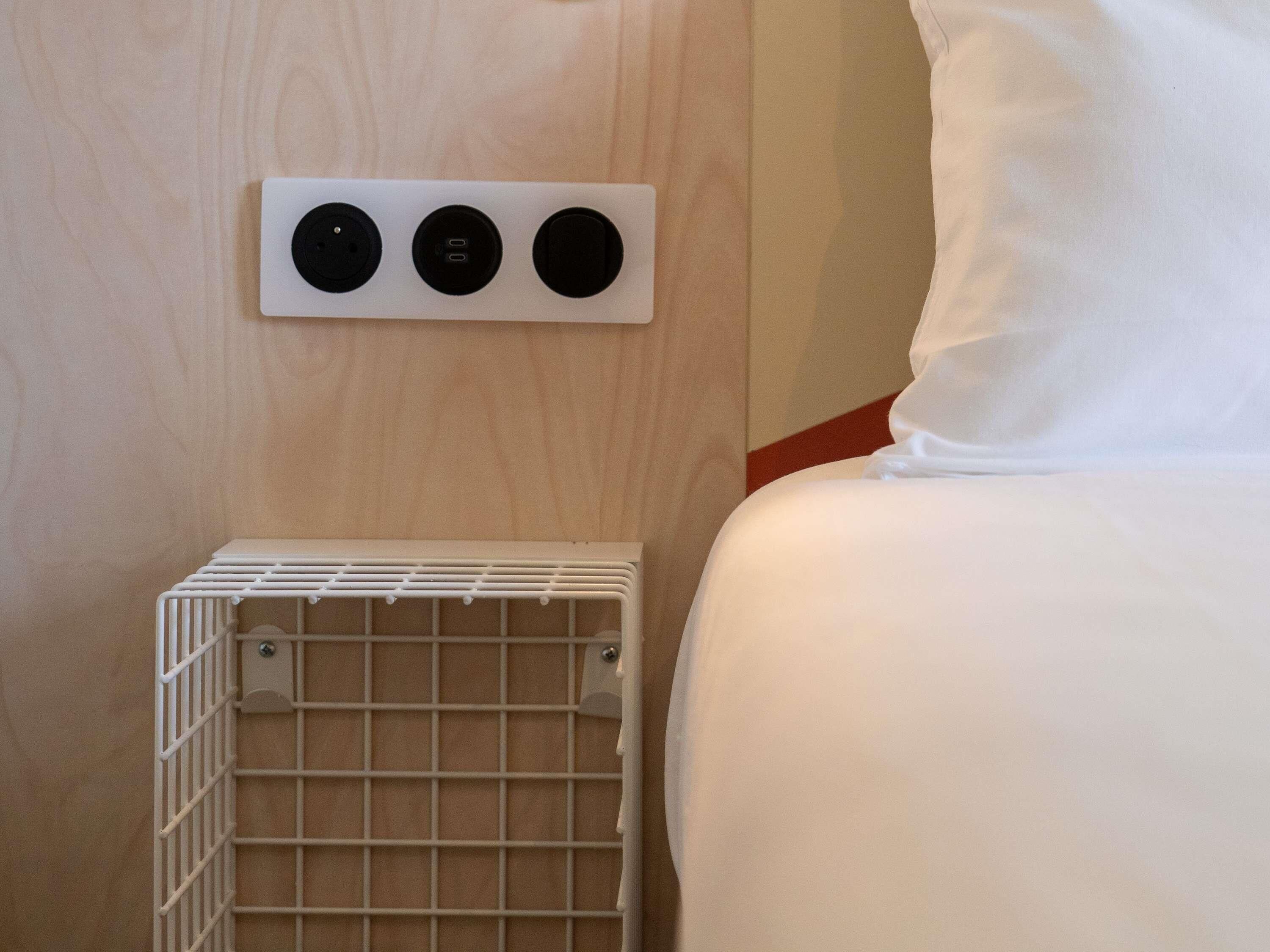 A hotel room with a bed, a white bedside table, and a wall-mounted electrical outlet with three sockets.