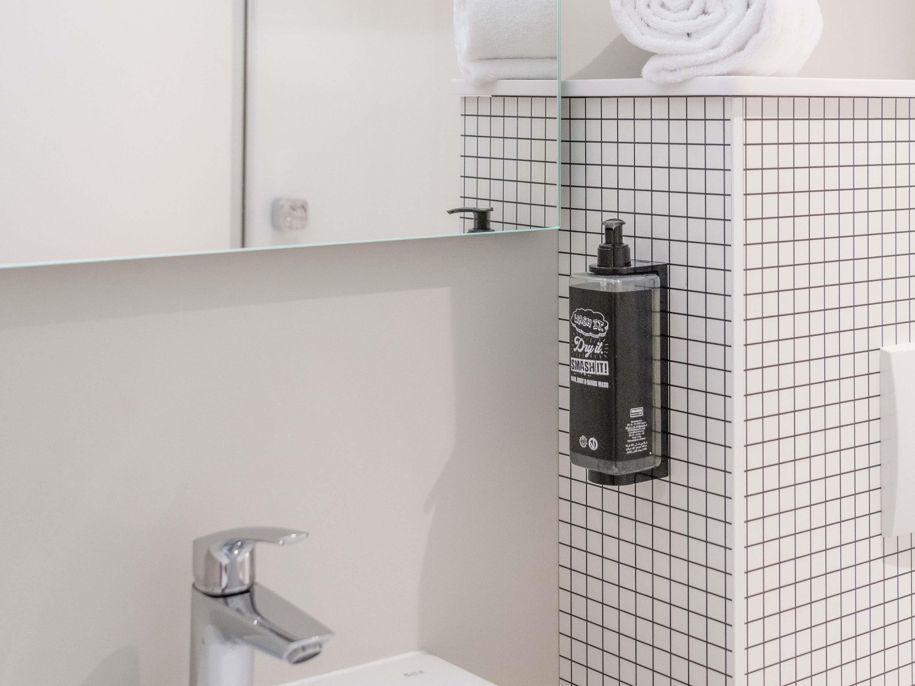 Un baño con pared de diseño cuadriculado, un perchero con una toalla enrollada, un dispensador de jabón y un lavamanos.
