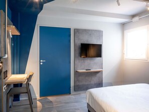 Moderni hotellihuone, jossa on sininen ovi, seinälle kiinnitetty televisio, pieni hylly, sänky ja työpöytä tuolilla.