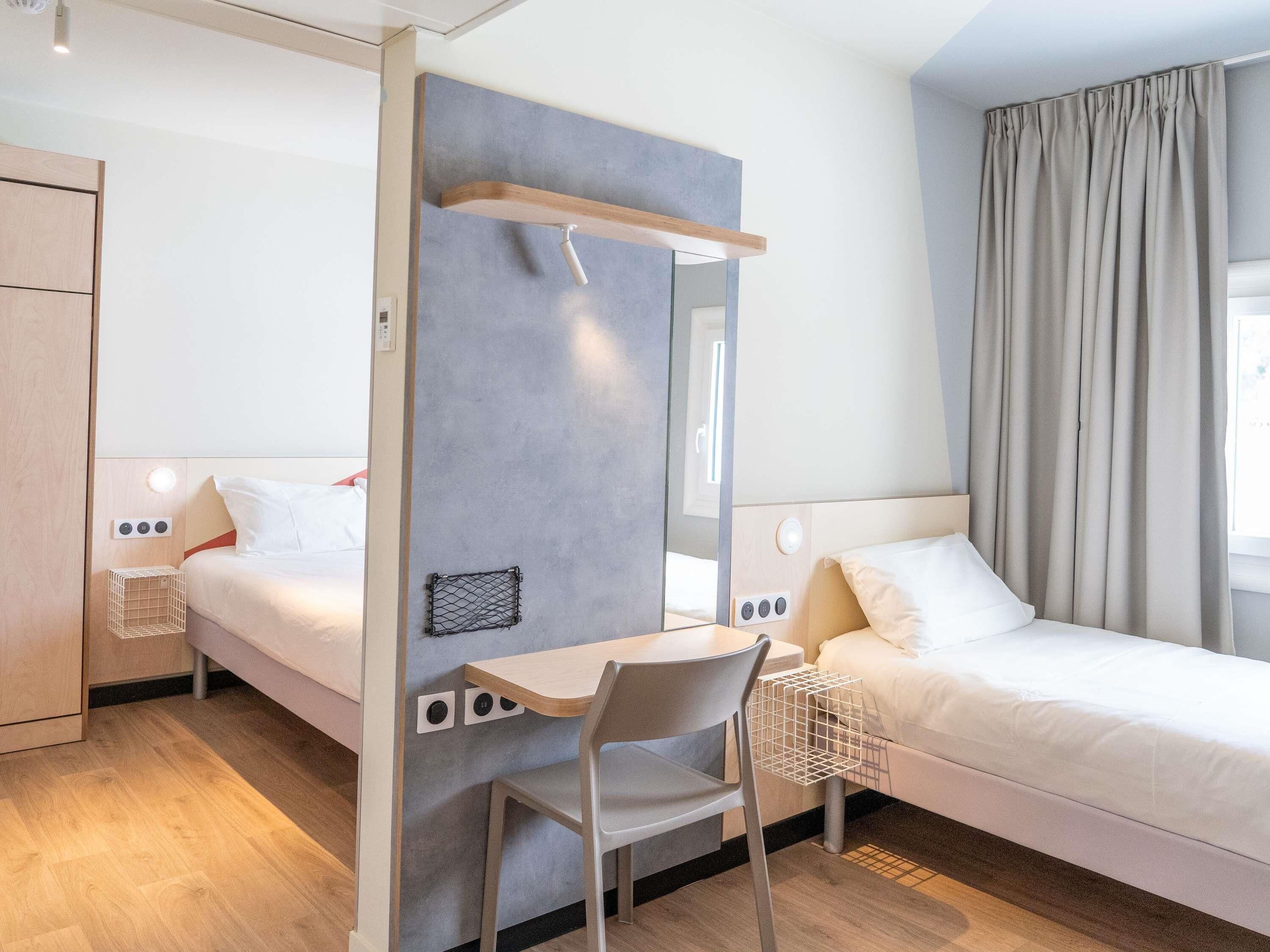 A modern hotel room with a bed, a desk with a chair, a mirror, and a wardrobe.