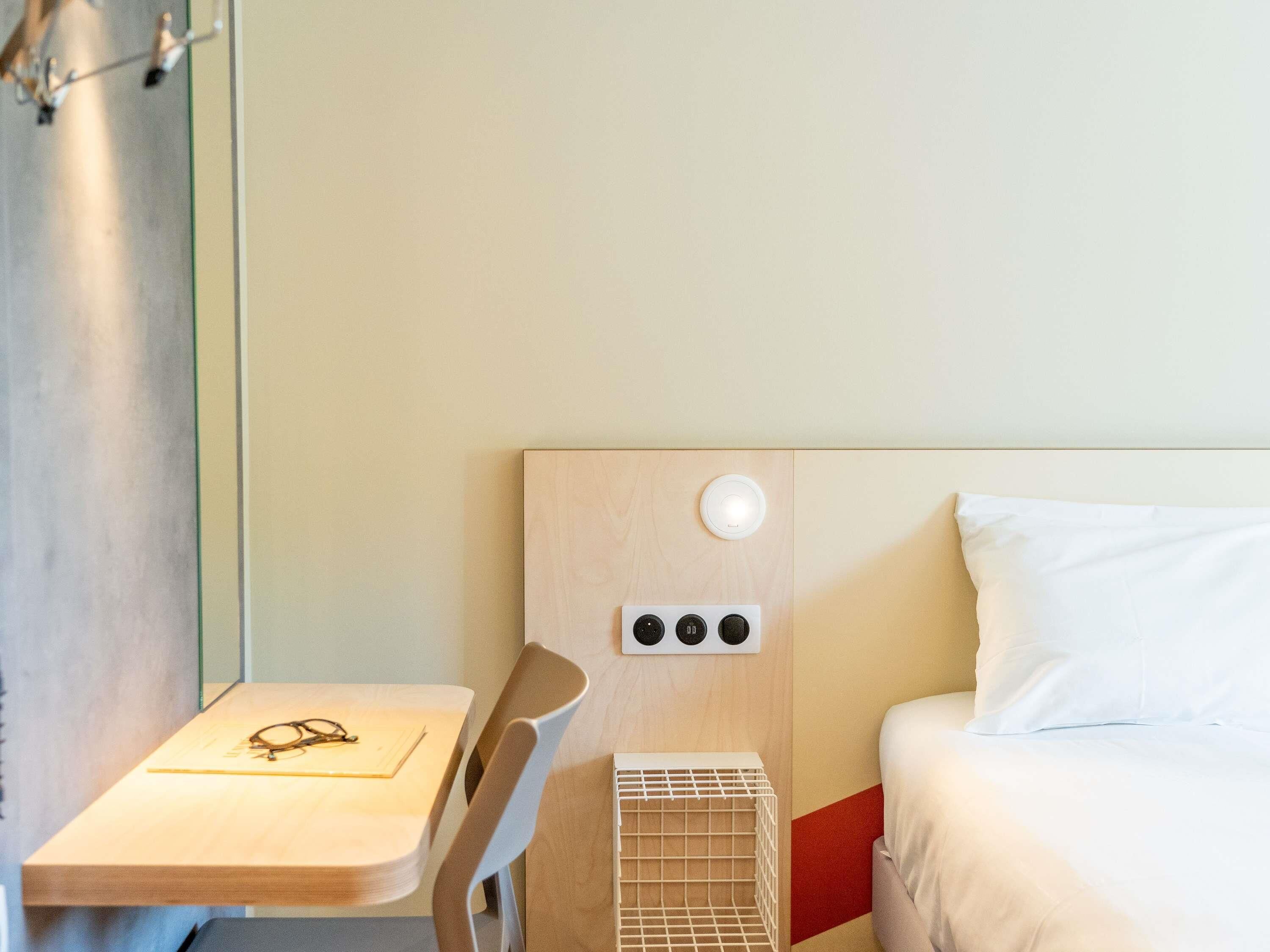Ett hotellrum med en säng, ett nattduksbord med glas och en anteckningsbok, en liten vit hylla och en väggmonterad kontrollpanel.
