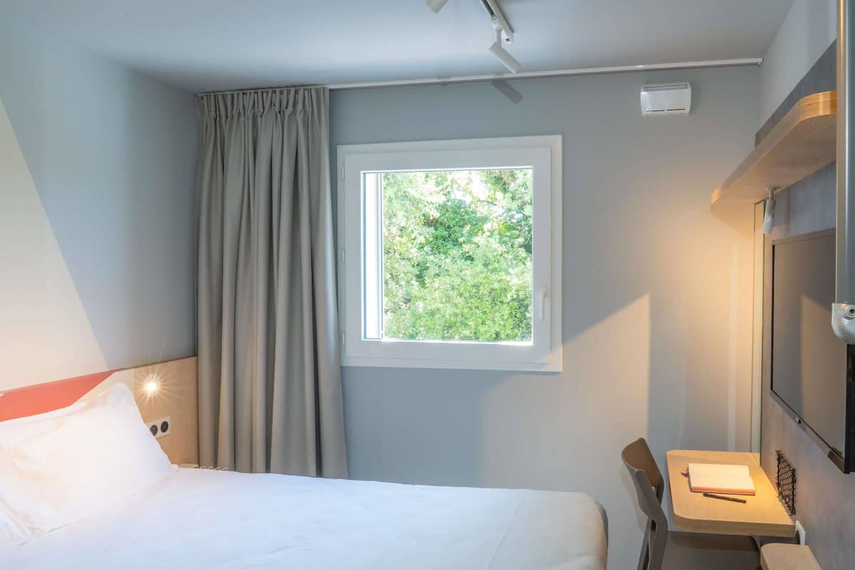 A hotel room with a bed, a desk, a chair, a window with a view of greenery, and a mounted TV.