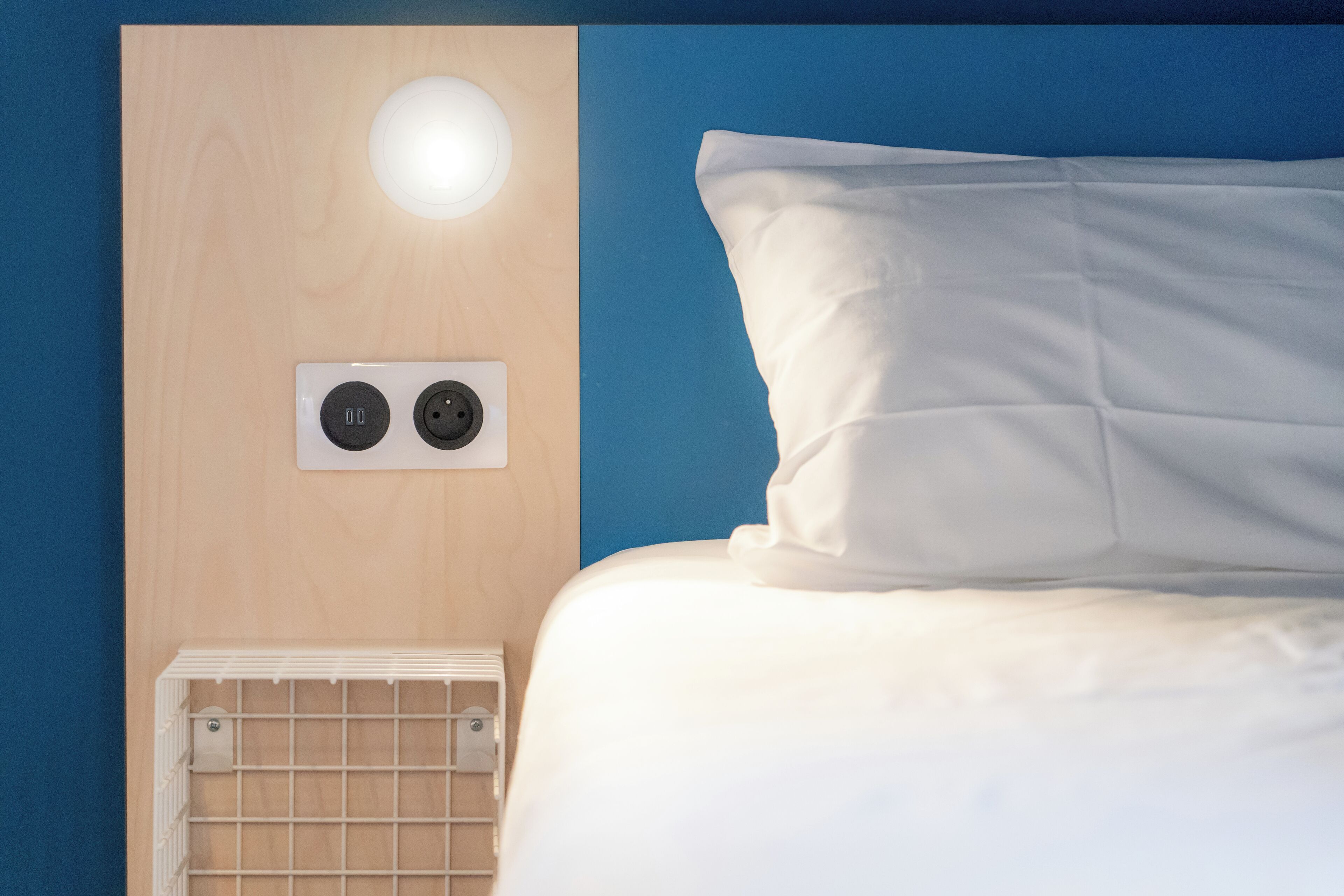 A bed with white bedding, a white wall with a light fixture, and a power outlet with two plugs.