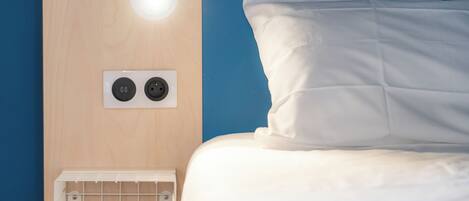 A bed with white bedding, a white wall with a light fixture, and a power outlet with two plugs.