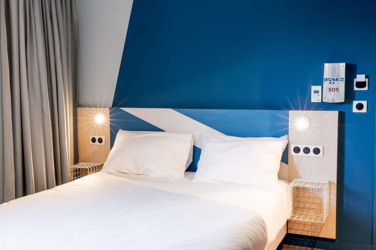 A modern hotel room with a large bed, two bedside tables, a wall-mounted SOS emergency phone, and a control panel with buttons.