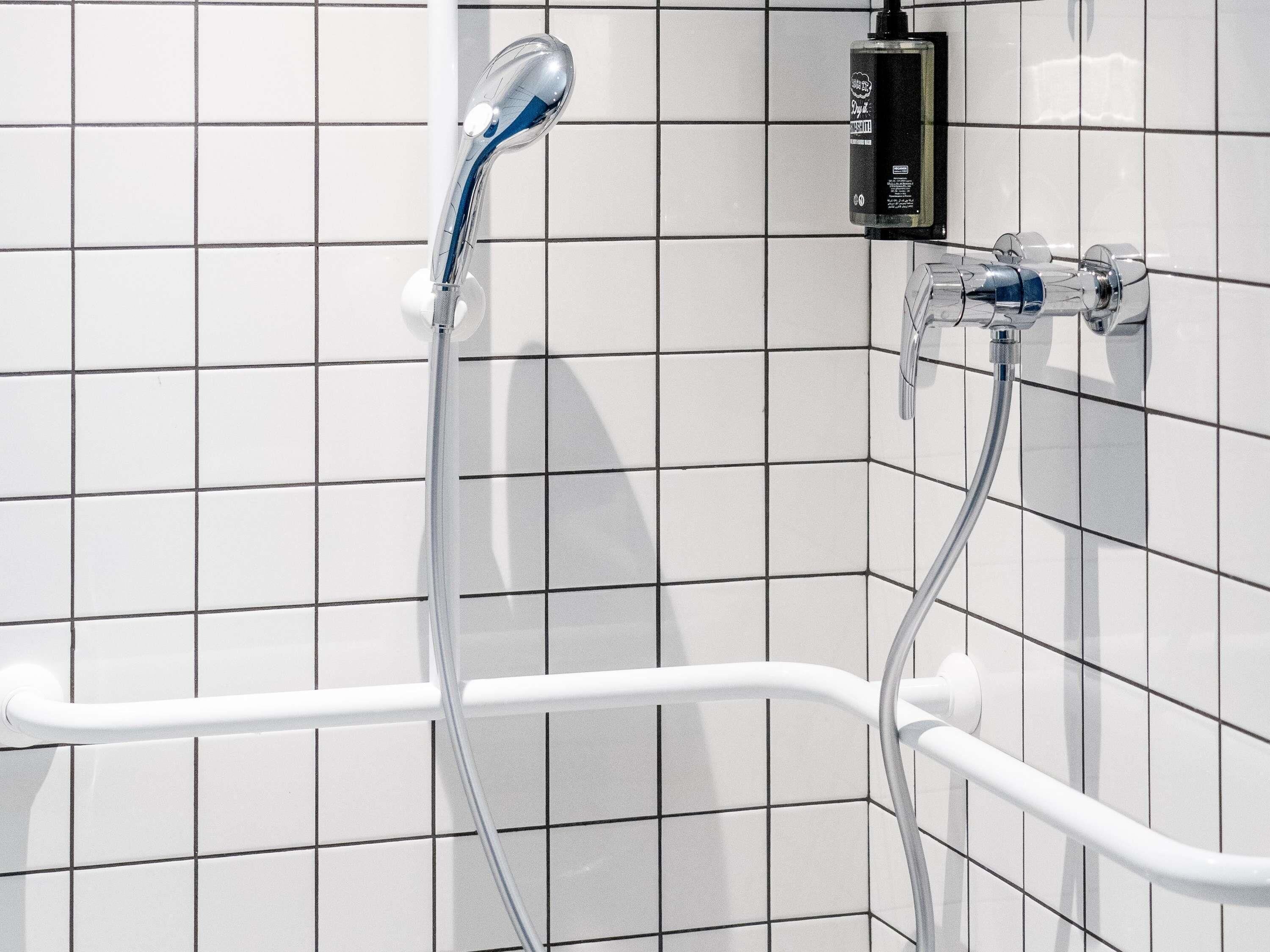 Un baño con paredes de azulejos blancos, una ducha con cabezal de ducha manual y un accesorio de ducha manual.