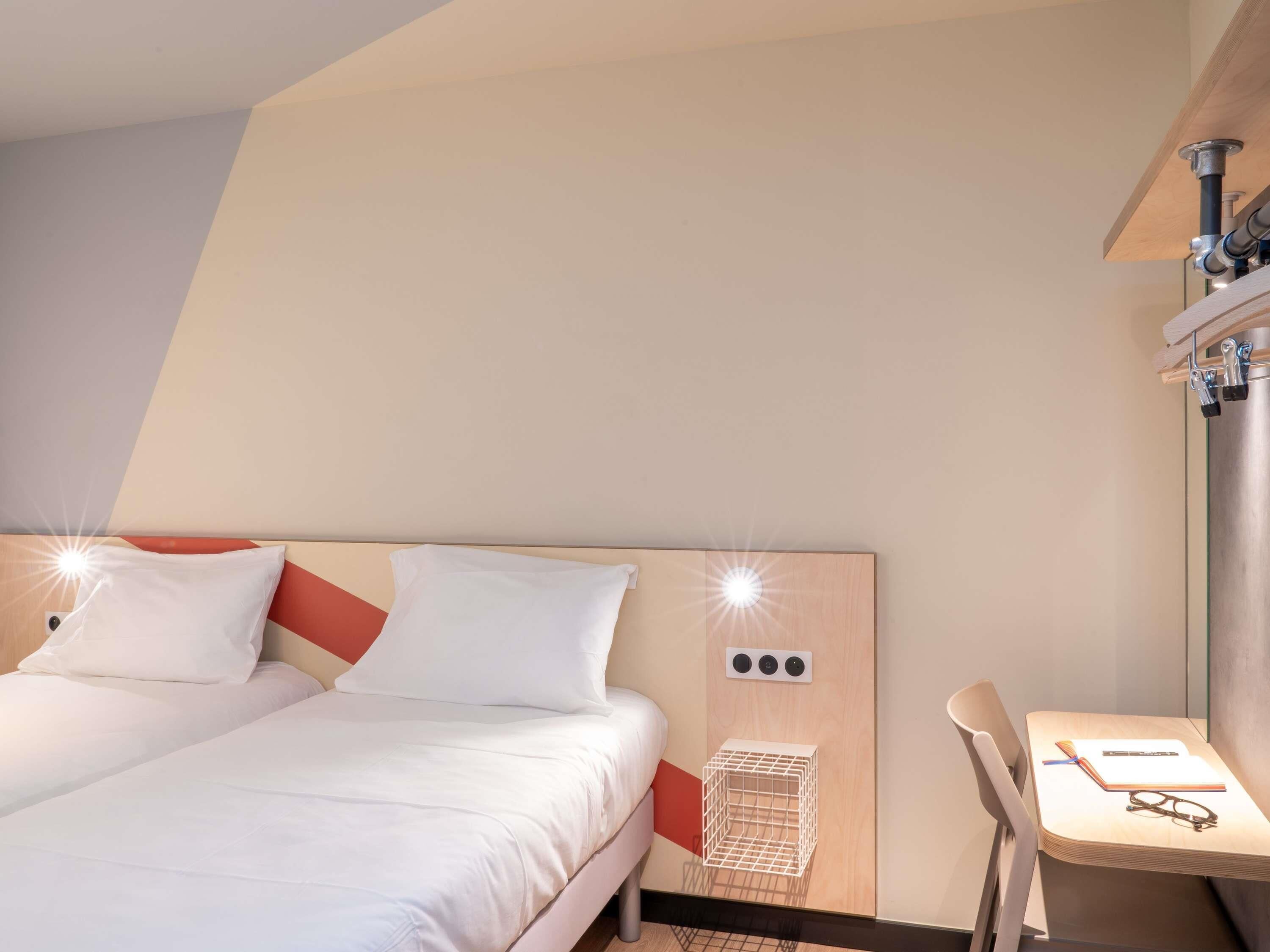 A hotel room with a bed, a desk with a chair, a small white shelf, and a wall-mounted shelf with a metal container.