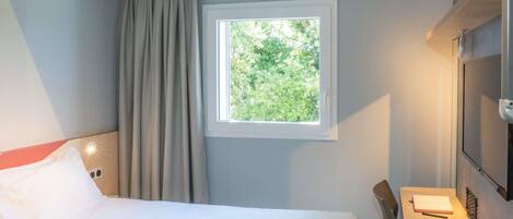 A hotel room with a bed, a desk, a chair, a window with a view of greenery, and a mounted TV.