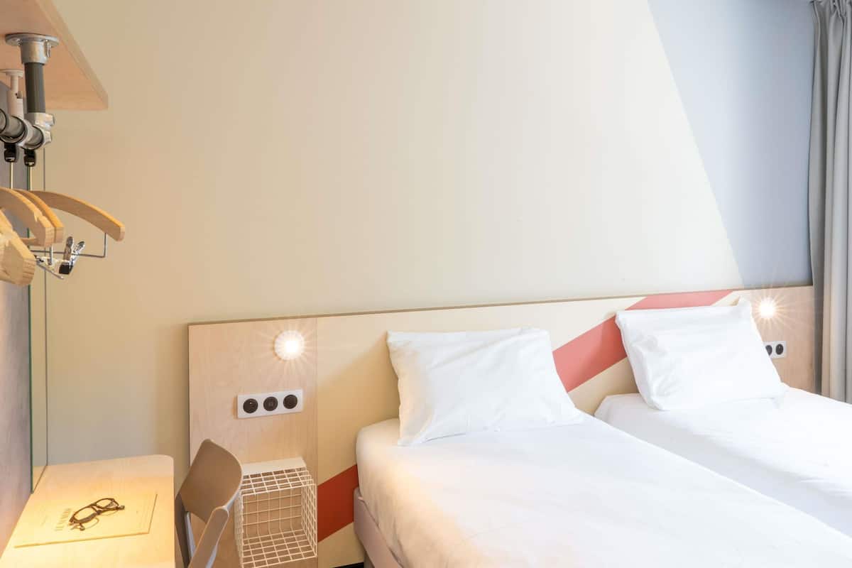 A hotel room with a bed, a chair, a table with glasses on it, and a wall-mounted lamp.