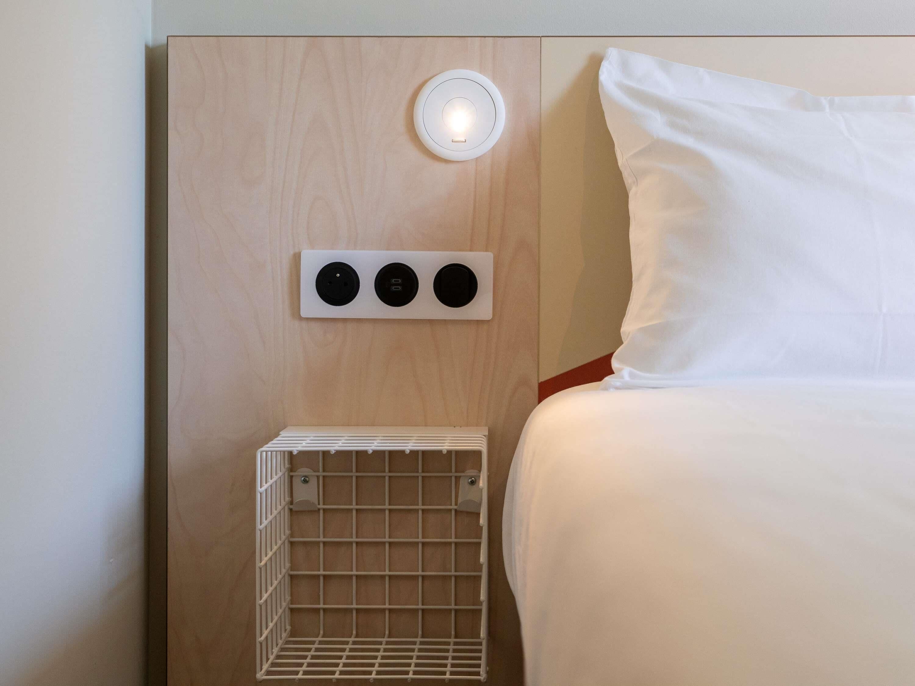 Habitación de hotel con cama, mesita de noche con lámpara y toma de corriente en la pared.