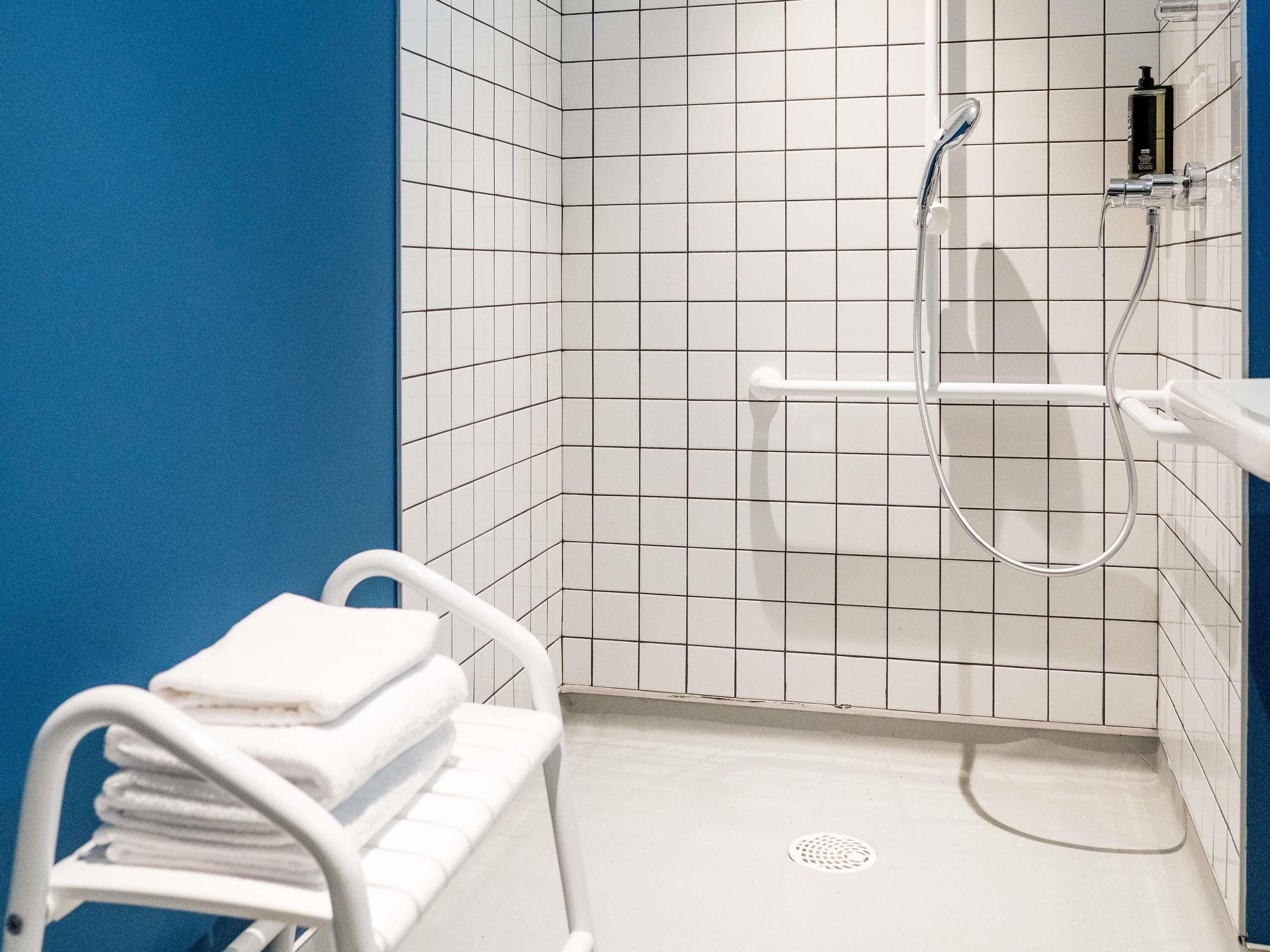 Een badkamer met witte betegelde wanden, een blauwe accentmuur, een witte douche met handdouche en een witte handdoekhouder met handdoeken.