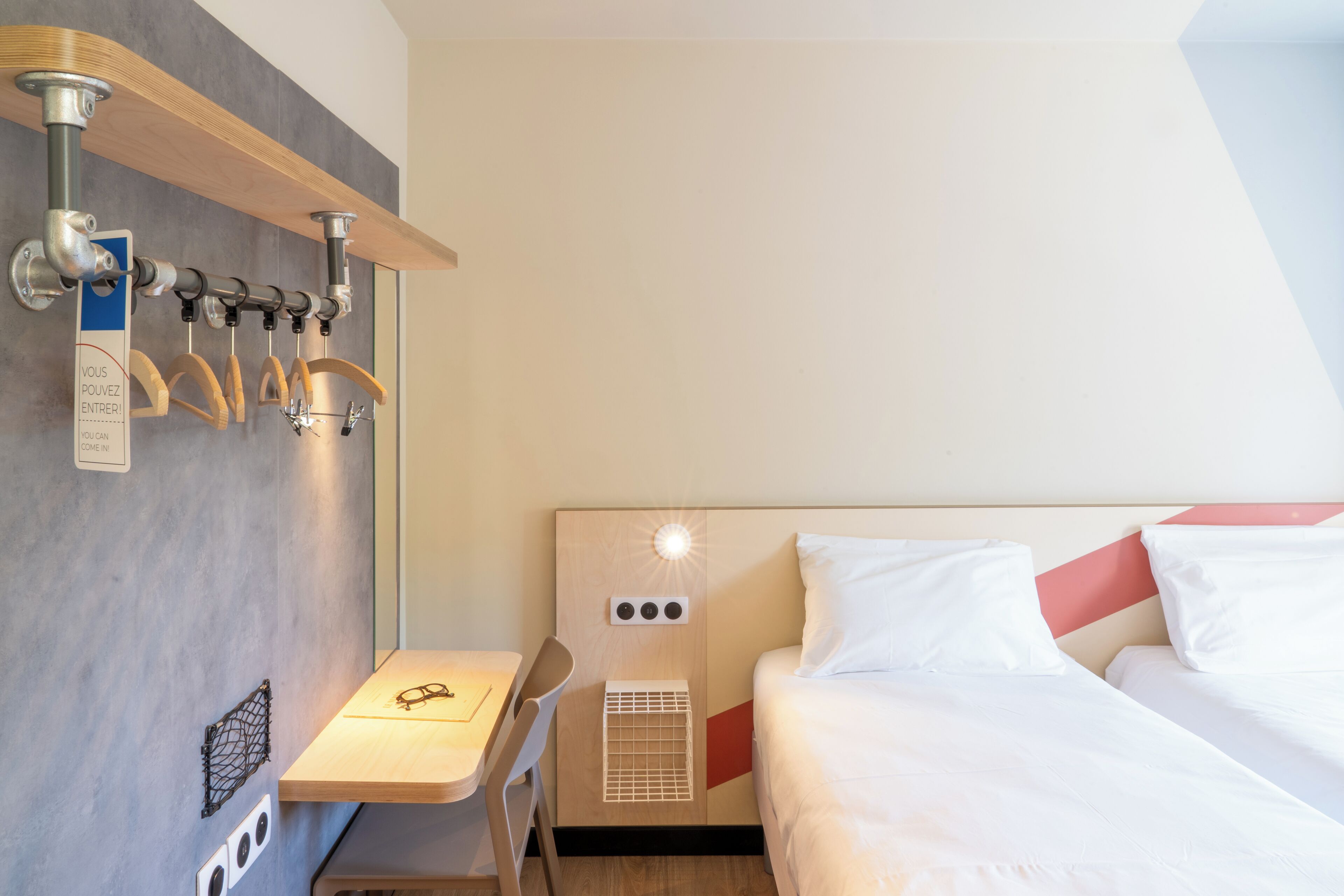 A hotel room with a bed, a desk, and a chair. There is a coat rack with multiple hooks.