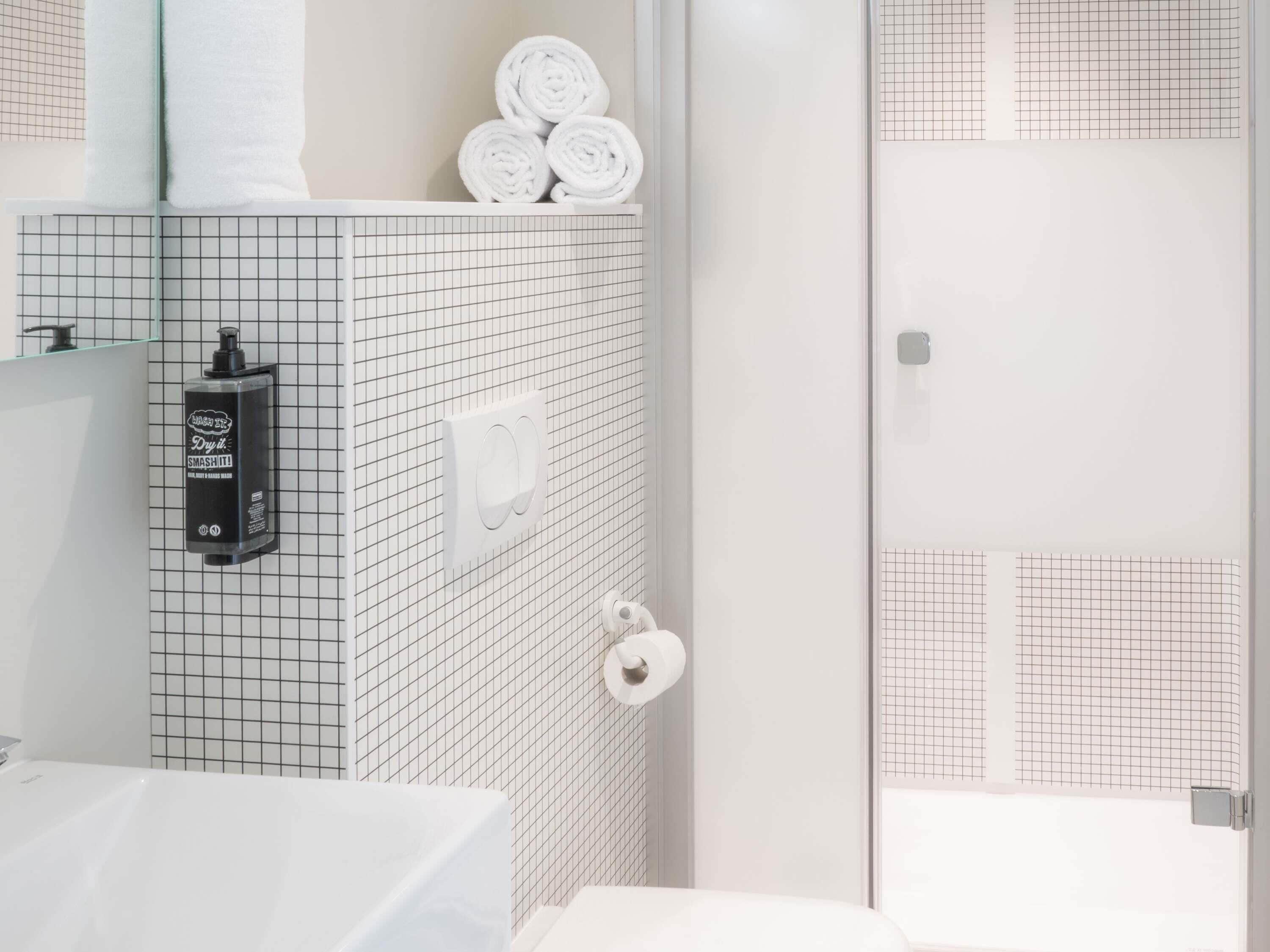 Ein Badezimmer mit einer Wandoberfläche im Gittermuster, einem Seifenspender, einem Handtuchhalter mit aufgerollten Handtüchern und einem Toilettenpapierhalter.