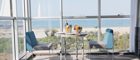 Habitación moderna con un ventanal grande con vistas a la playa, dos sillas, una mesita con una botella y vasos, y una vista del océano y edificios lejanos.