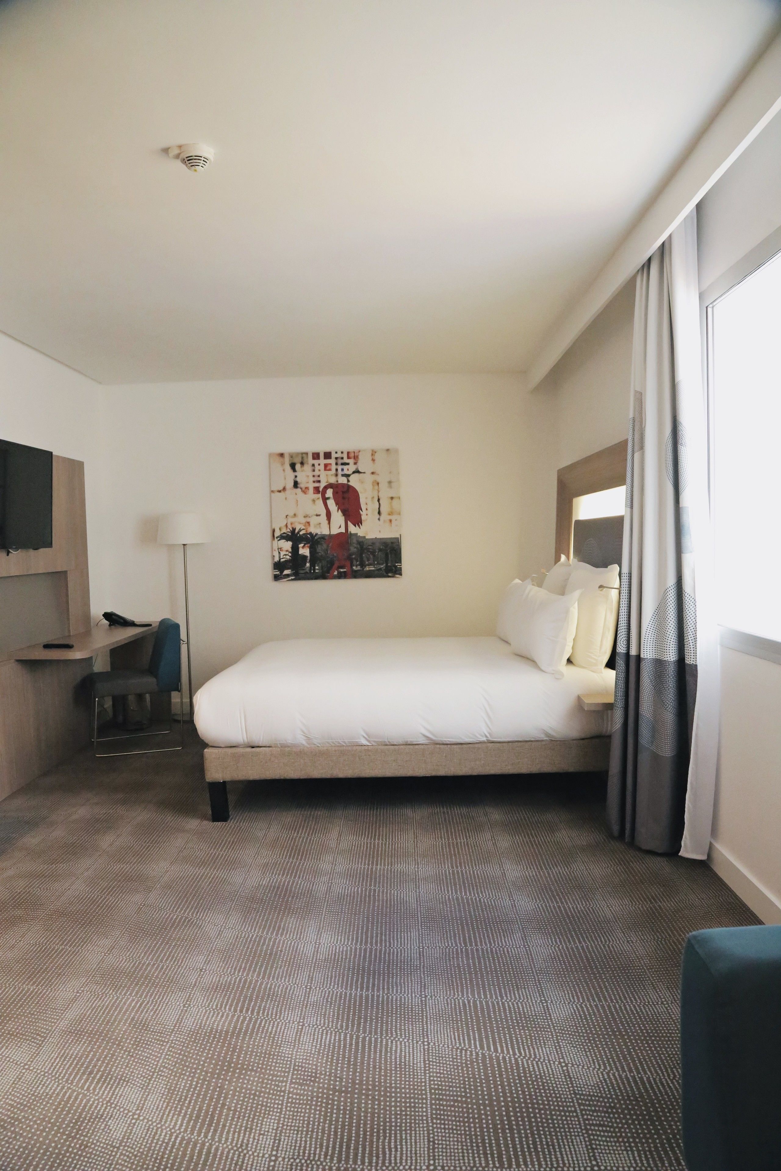 Una habitación de hotel con cama, escritorio, silla, lámpara y un cuadro en la pared.