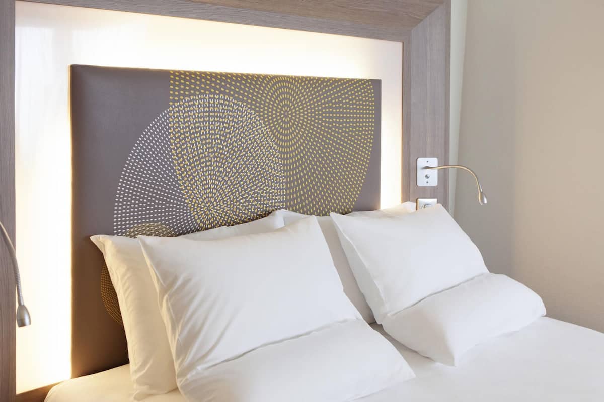 Una habitación de hotel con cama, ropa de cama blanca, cabecero con diseño geométrico y una lámpara de noche.