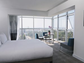 Habitación de hotel con una cama grande, una mesita pequeña, dos sillas y vistas a la ciudad a través de amplios ventanales.
