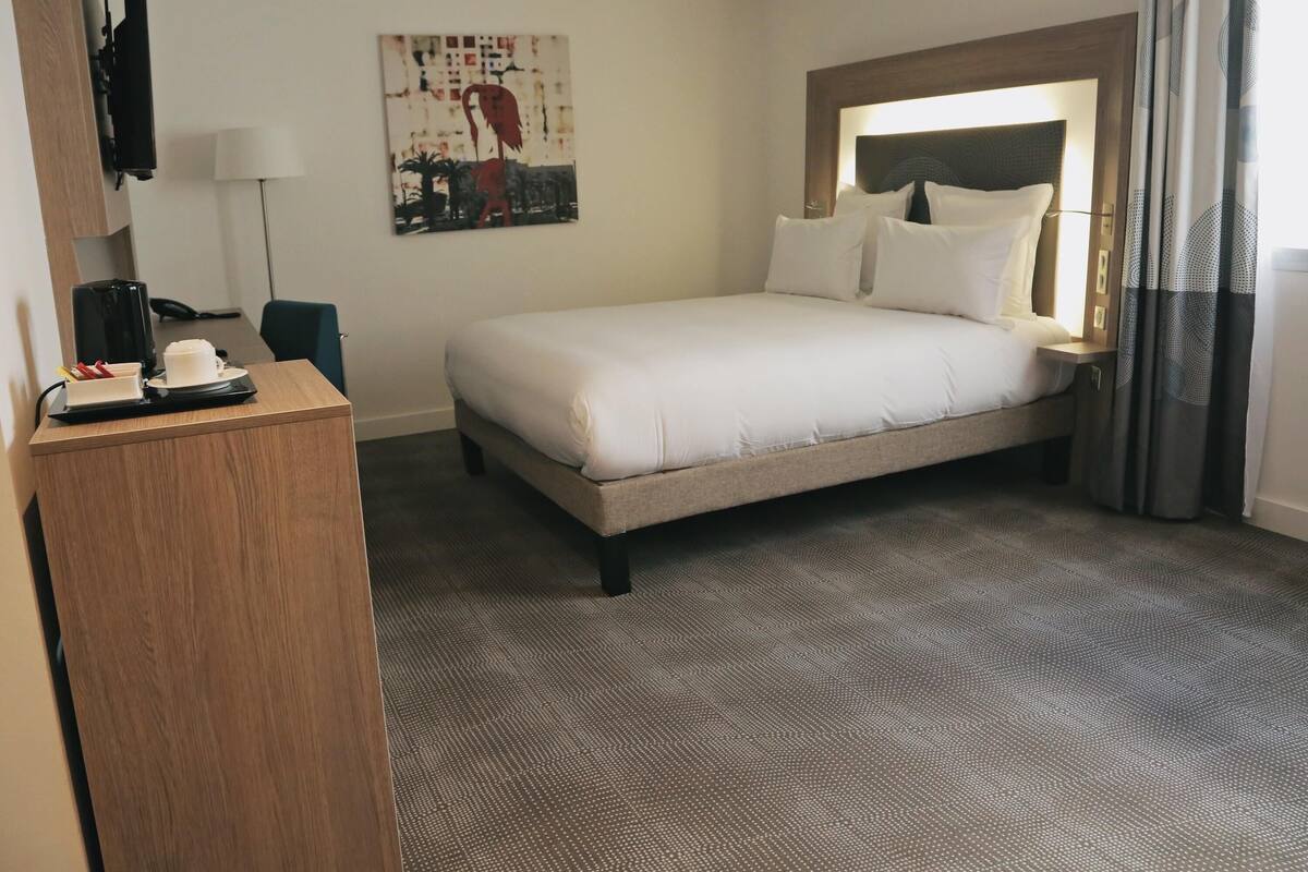 A hotel room with a bed, a desk with a phone and a coffee maker, a painting on the wall, and a window with curtains.