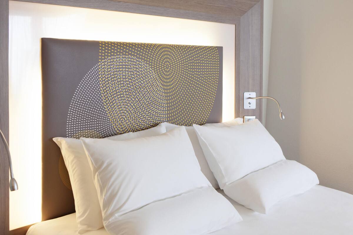 A hotel room with a bed, white bedding, a headboard with a geometric pattern, and a bedside lamp.