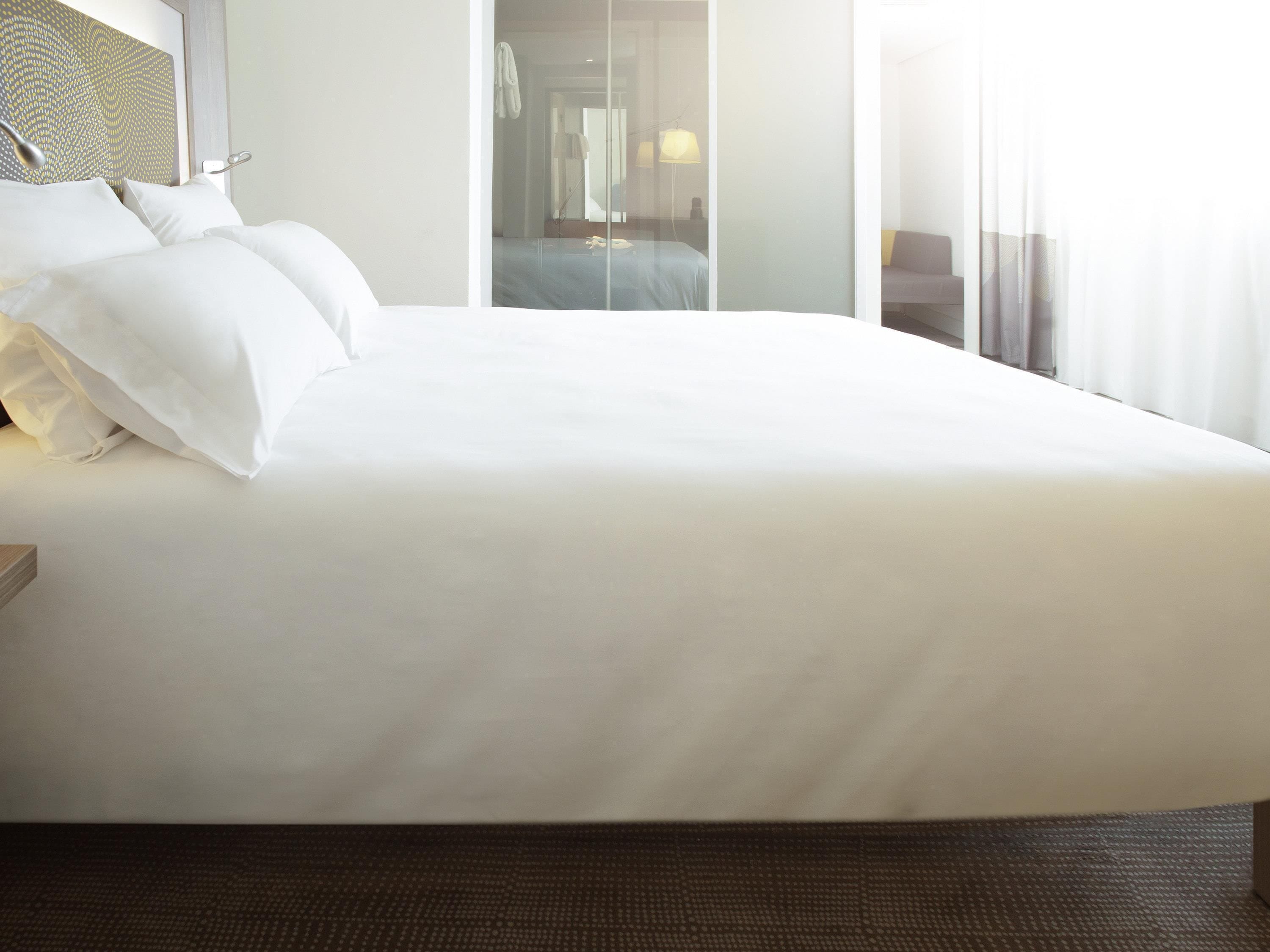 Una cama grande con sábanas blancas, cabecera con estampado geométrico, y vista a un pasillo con puerta y lámpara.