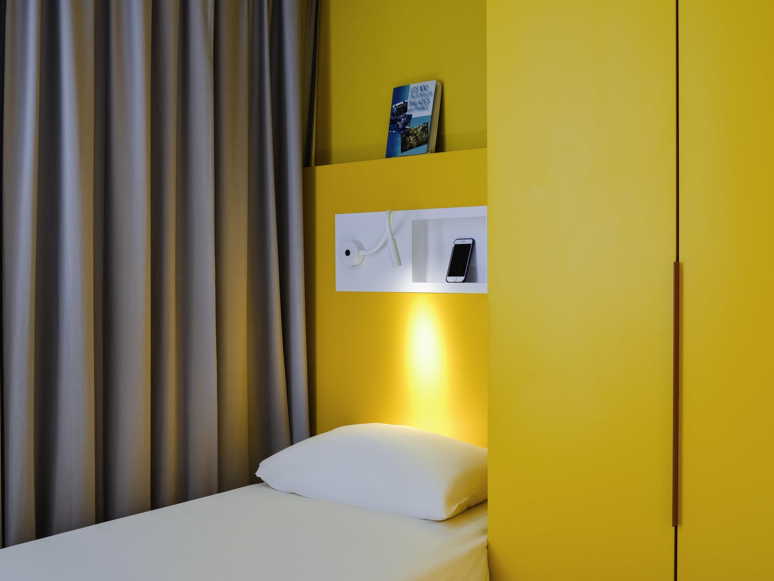A hotel room with a bed, a white pillow, a nightstand with a phone, a book on the nightstand, and a curtain.
