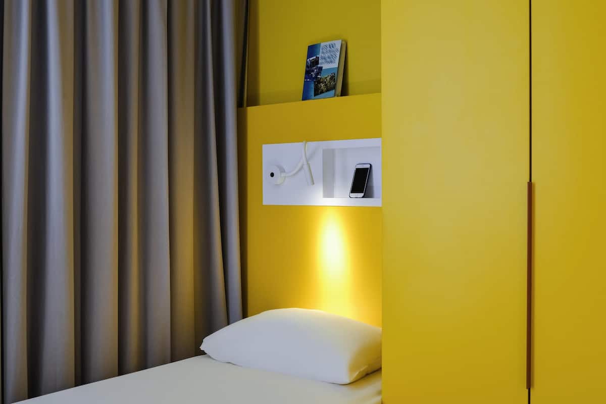 Quarto de hotel com cama, travesseiro branco, mesa de cabeceira com telefone, um livro na mesa de cabeceira e cortina.