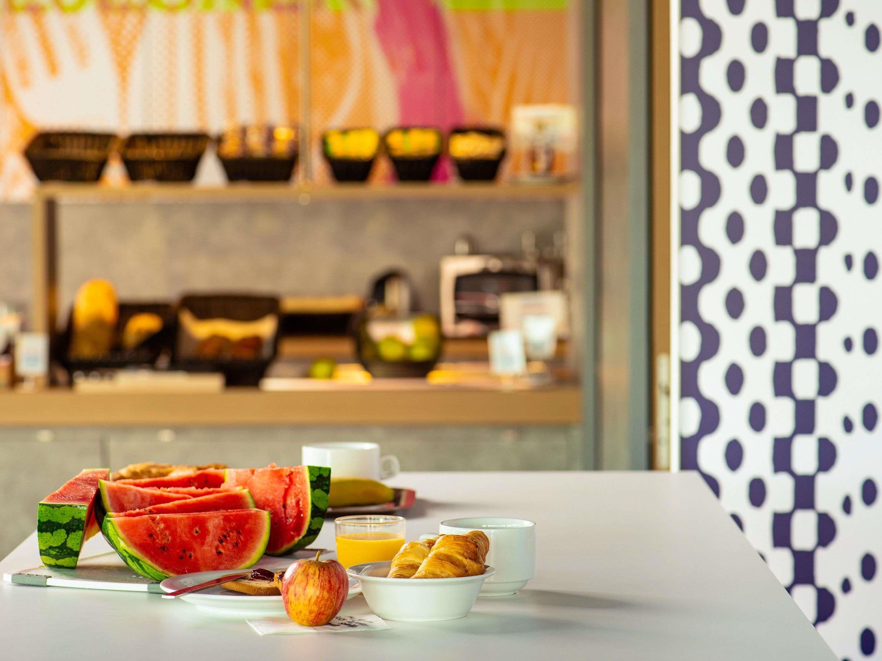A breakfast setting with watermelon, apple, orange juice, croissants, and coffee cups on a table.