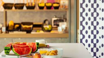 A breakfast setting with watermelon, apple, orange juice, croissants, and coffee cups on a table.