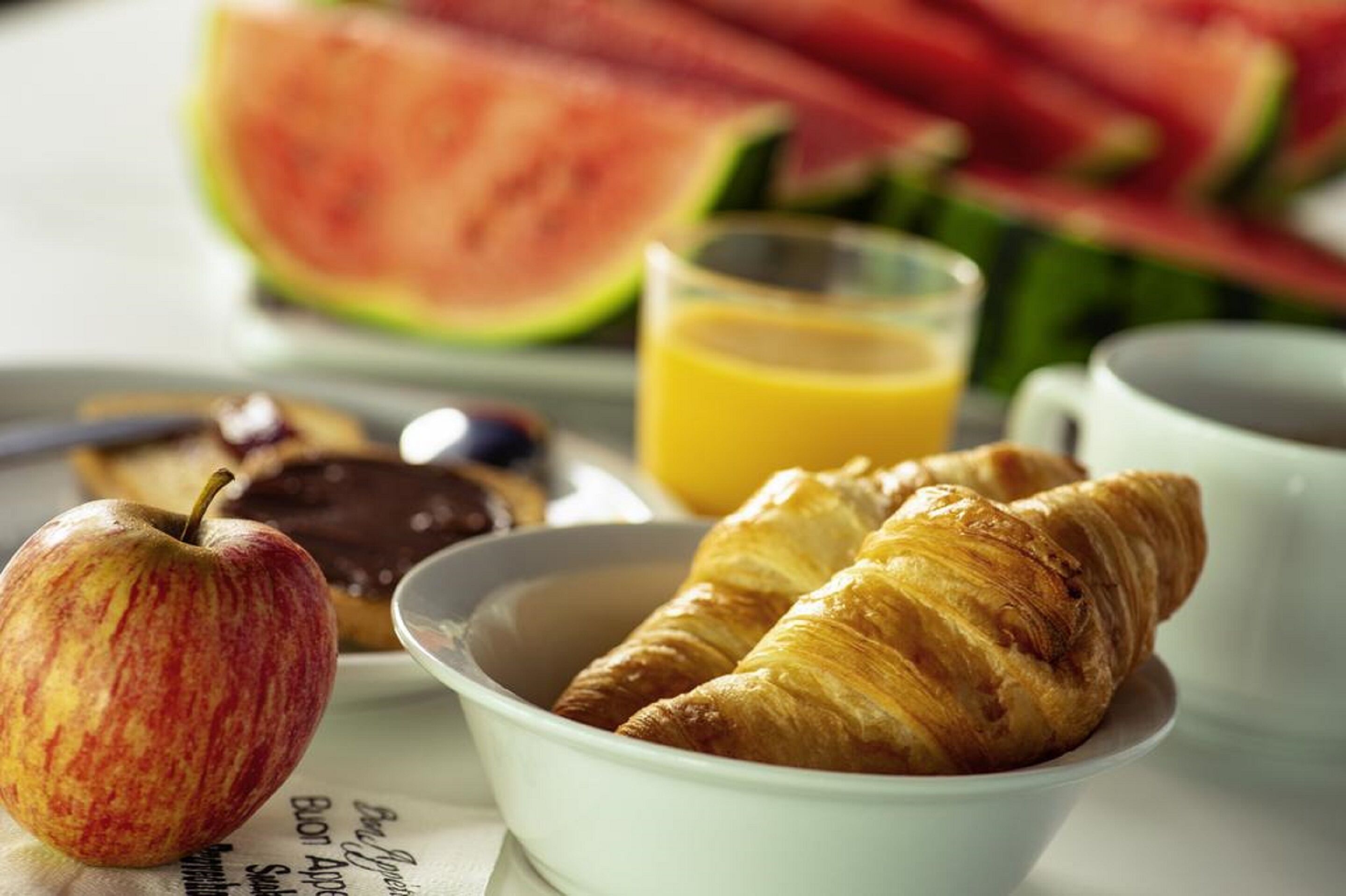 Un desayuno con croissants, una manzana, un vaso de zumo de naranja y una porción de sandía.